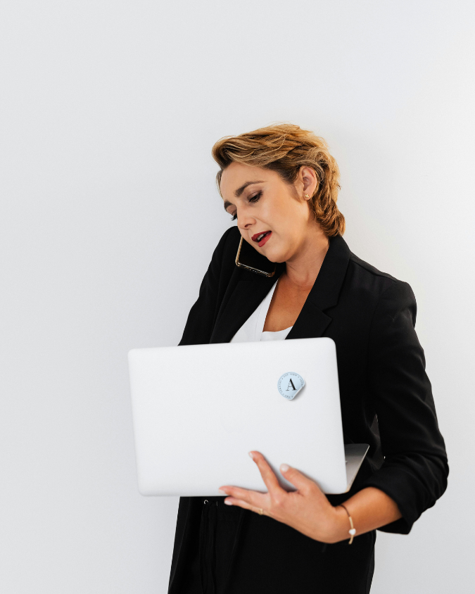 Inhyrd ekonomichef som arbetar med ekonomistyrning. En kvinna i svart kavaj håller en laptop medan hon pratar i telefon med hörlurar.