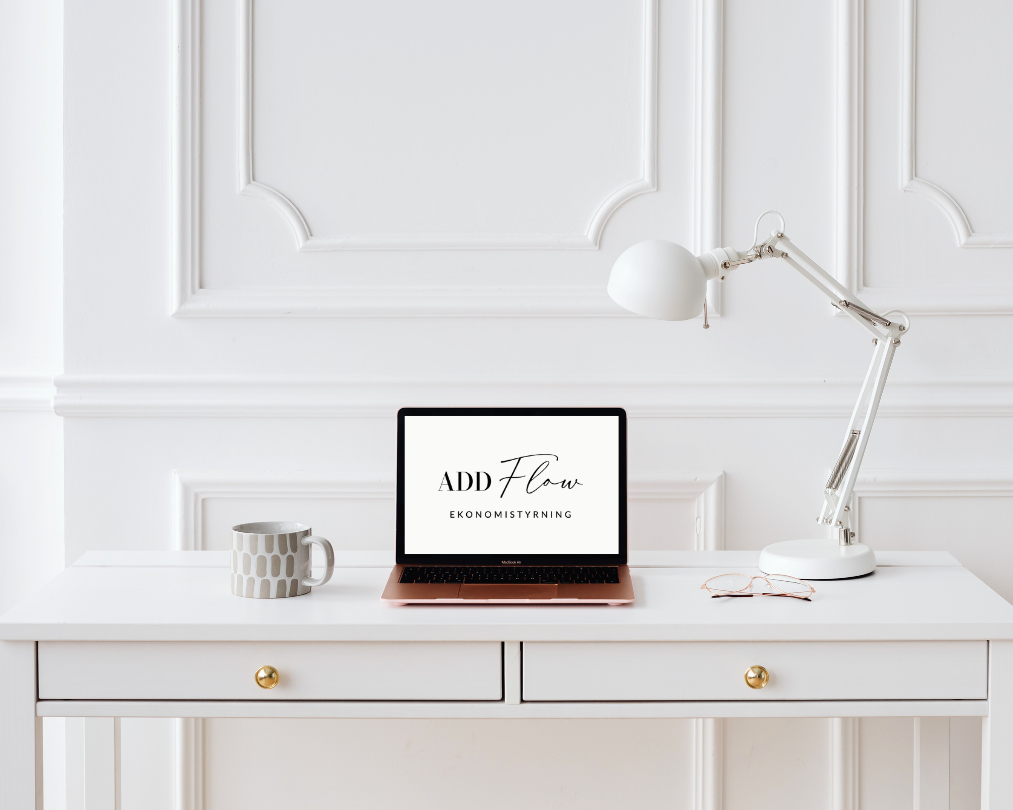 Ett skrivbord med en bärbar dator, ett mugg, ett par glasögon och ett skrivbordslampa i ett vitt rum med väggpanelen. På datorskärmen visas Add Flows logotyp med texten ekonomistyrning under.