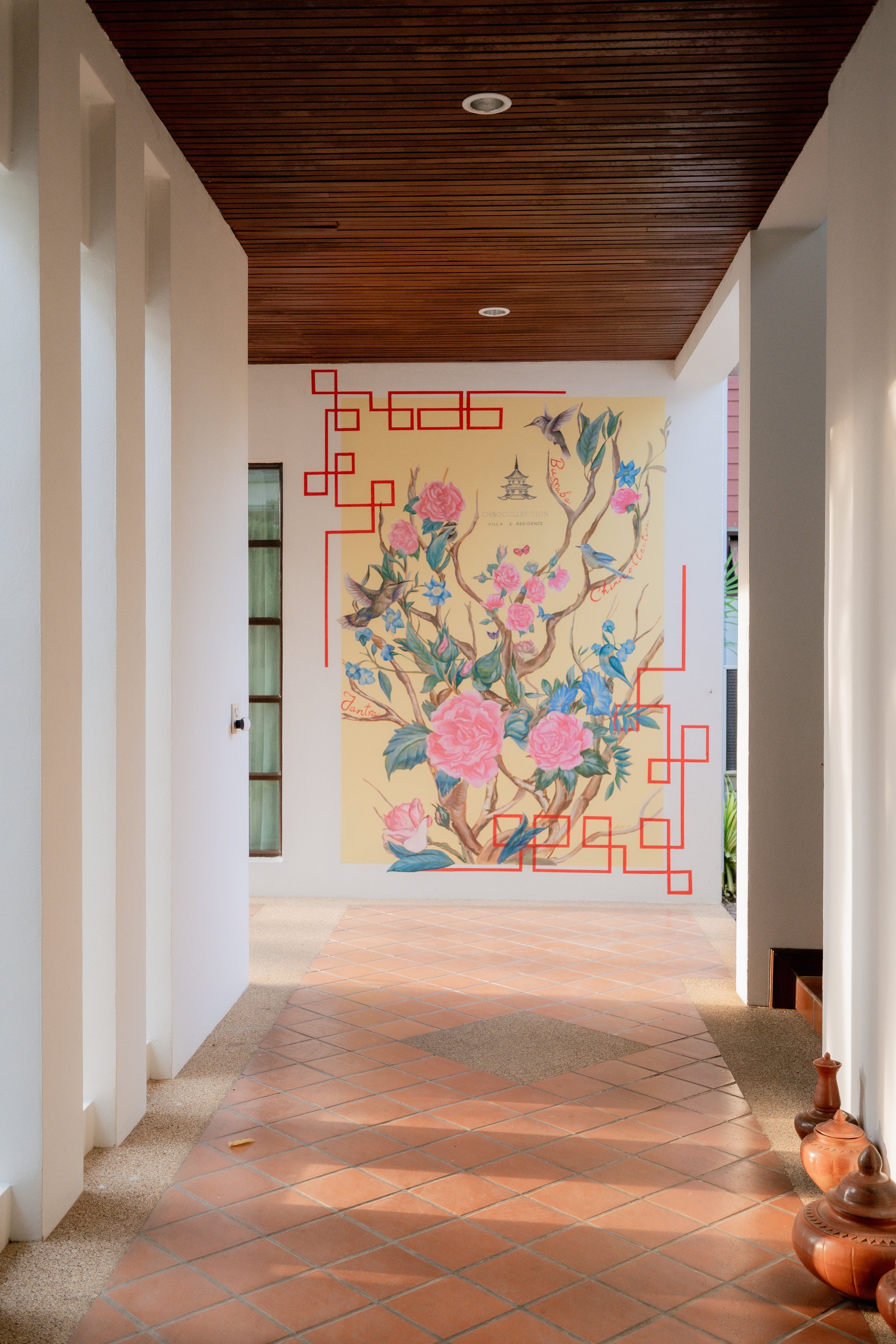 Interior hallway with a large mural of pink roses on a branch with blue flowers and hummingbirds, framed with red geometric lines, on a light yellow wall. Terracotta tiled floor and white walls, with a wooden ceiling and decorative pottery on the flo