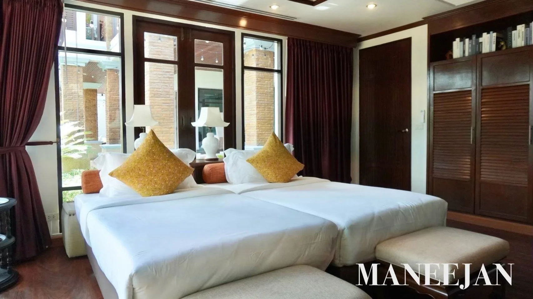 Hotel room with two beds, white linens, golden pillows, and a bedside lamp. Large windows with dark curtains and a view of a brick building and greenery outside. Wooden furniture and built-in bookshelves.