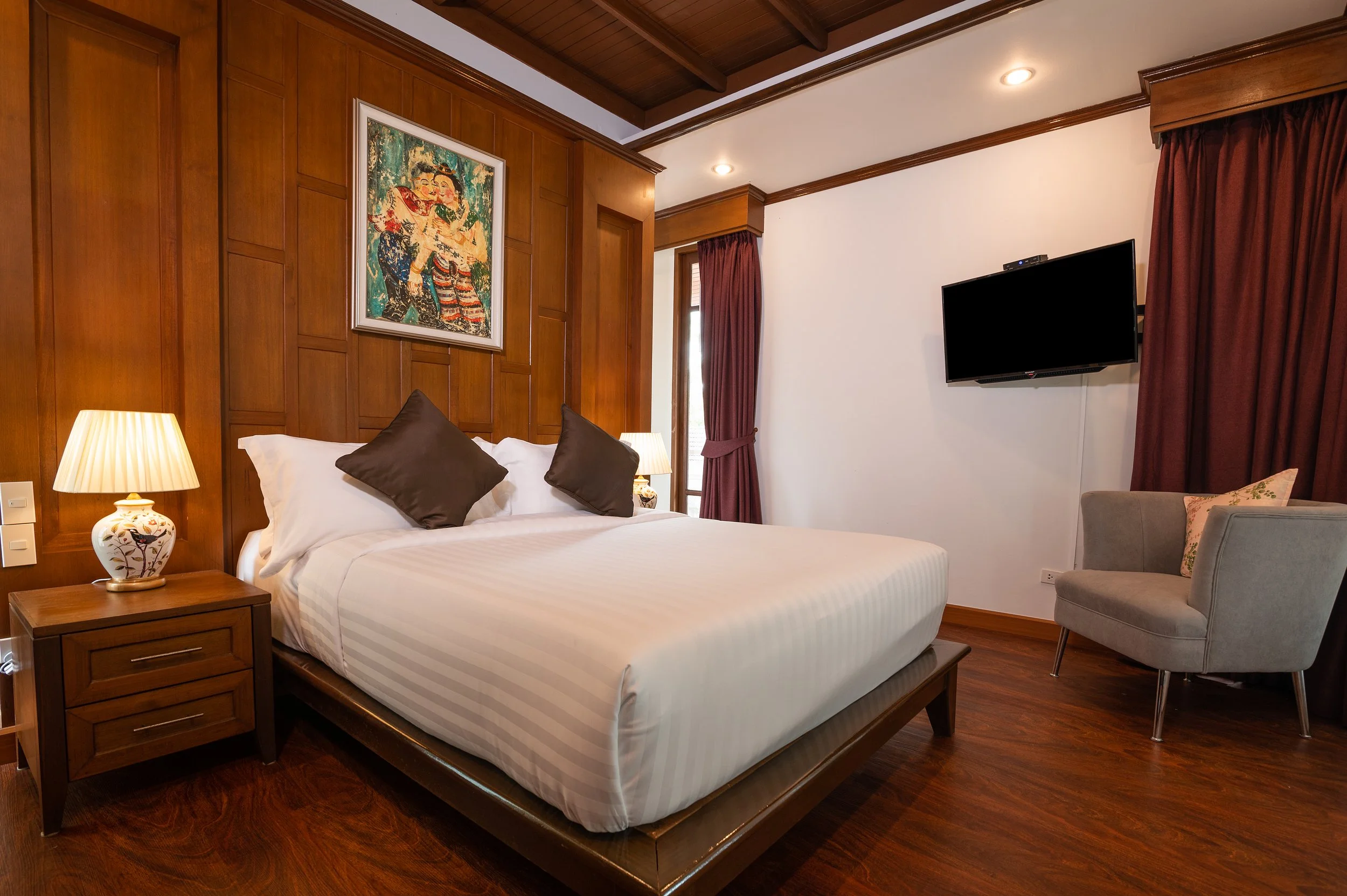Villa bedroom with wooden wall paneling, a bed with white linens and dark pillows, a bedside table with a lamp, a gray armchair, a wall-mounted TV, and purple curtains.
