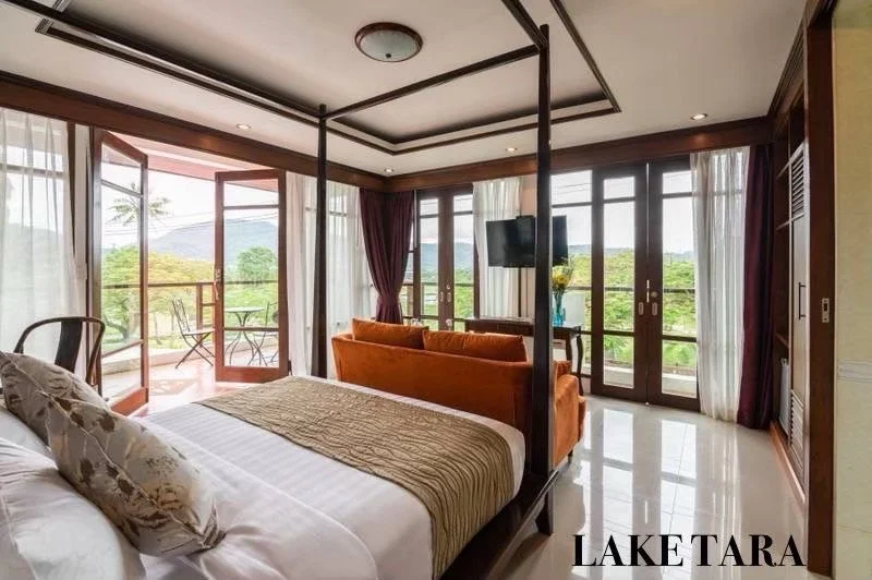 A bedroom with a four-poster bed, orange sofa, and large windows and balcony doors with curtains, overlooking green trees and mountains. Best large group villa Phuket