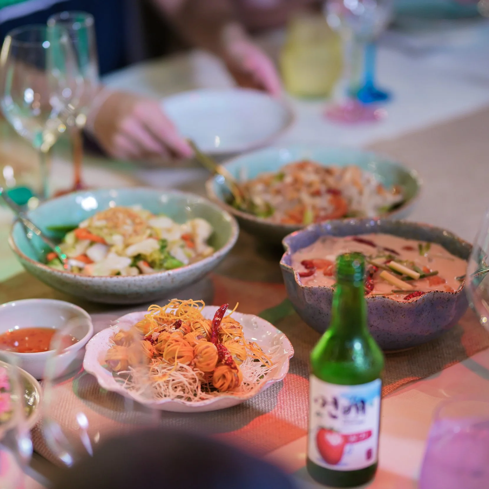 A table set with various bowls of colorful salads and dishes, a green bottle, and wine glasses, with a person in the background reaching for food. Private dining best villa Phuket