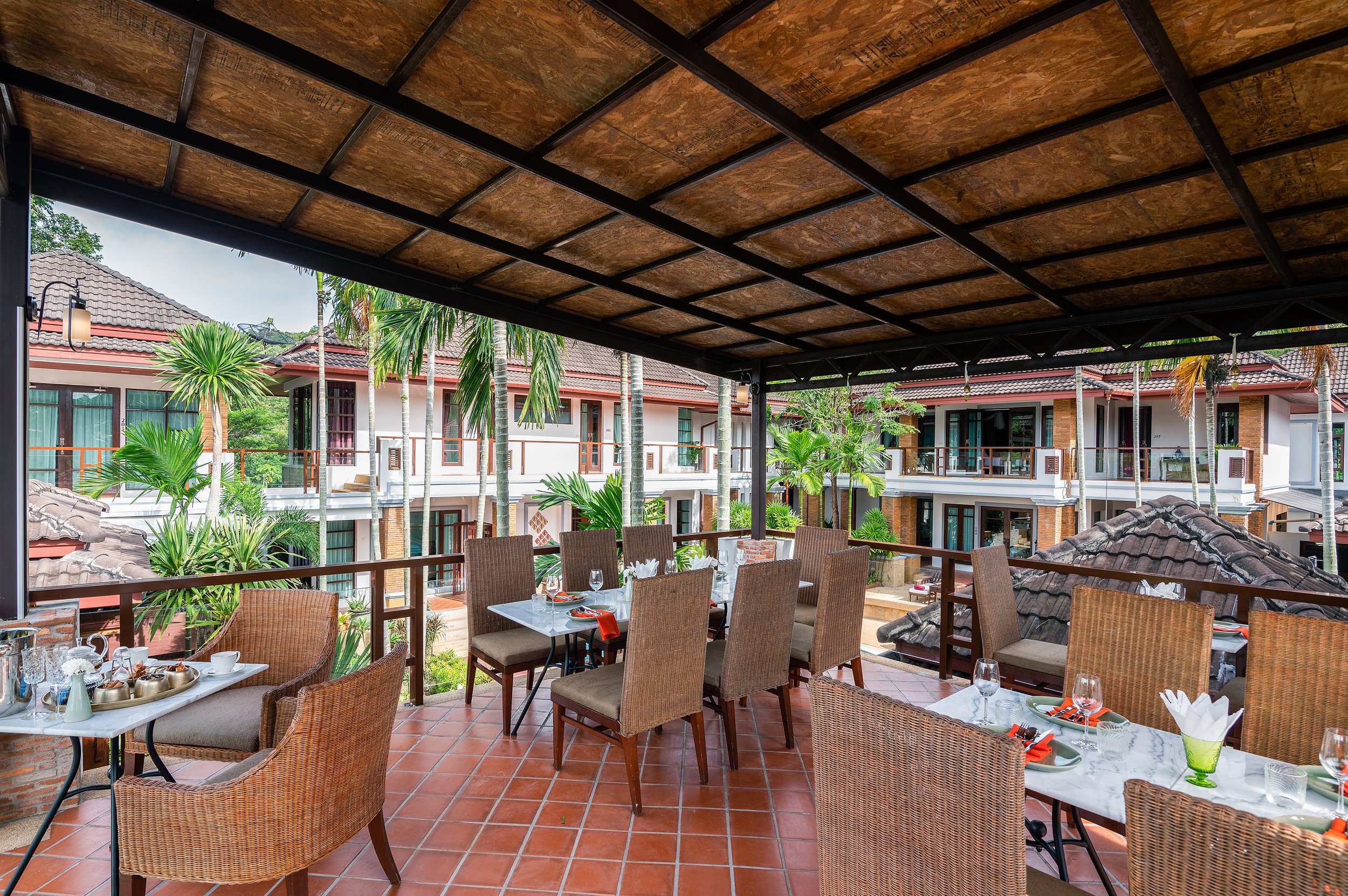 Outdoor restaurant patio with dining tables set for a meal, surrounded by tropical plants and neighboring buildings with balconies, under a wooden ceiling.