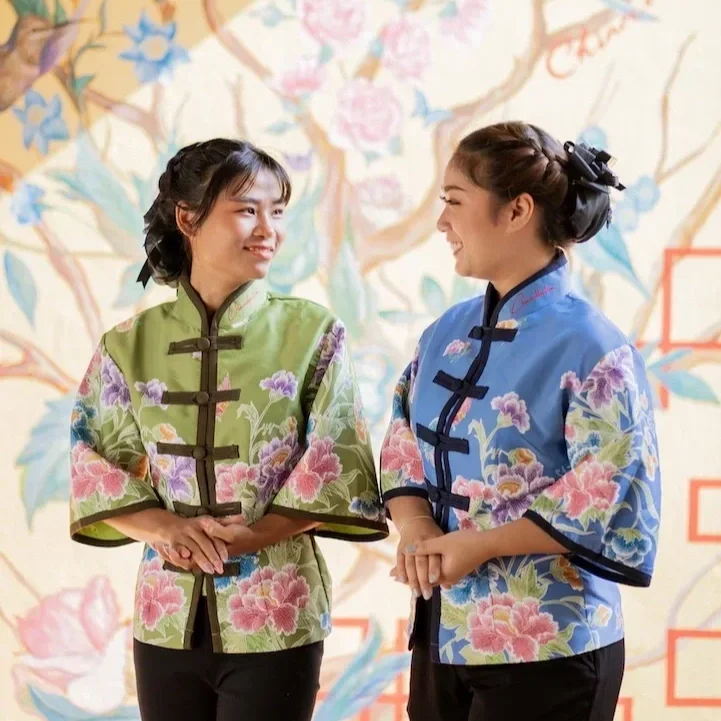 Two women in traditional Chinese cheongsam-style blouses with floral patterns, smiling and talking in front of a floral background. Spotlight service