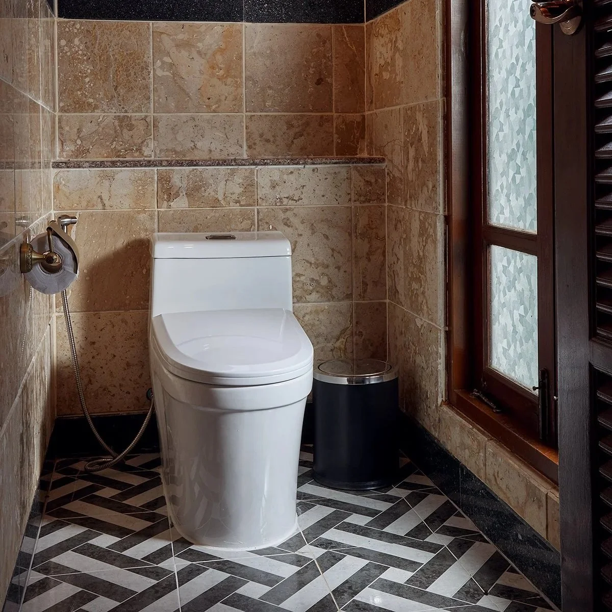A modern bathroom with a white toilet, a black trash bin, and a wooden framed window with frosted glass. The walls are tiled with beige and brown stone tiles, and the floor features a black and white geometric tile pattern.