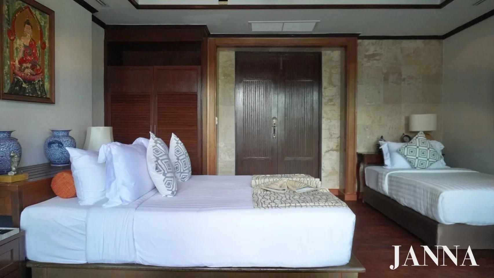 A hotel room with two beds, decorative pillows, lamps, and artwork. The room features wood accents and stone wall elements.