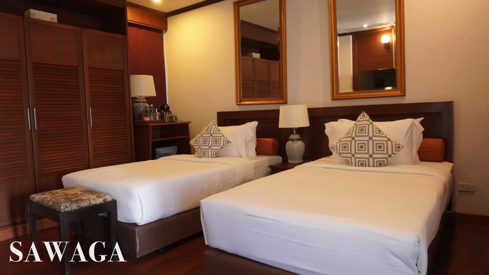 Hotel room with two single beds, decorative pillows, wooden headboards, double mirrors above the beds, nightstand with lamp, built-in wooden wardrobe, and a small table with a cushion, warm lighting, and wood flooring.