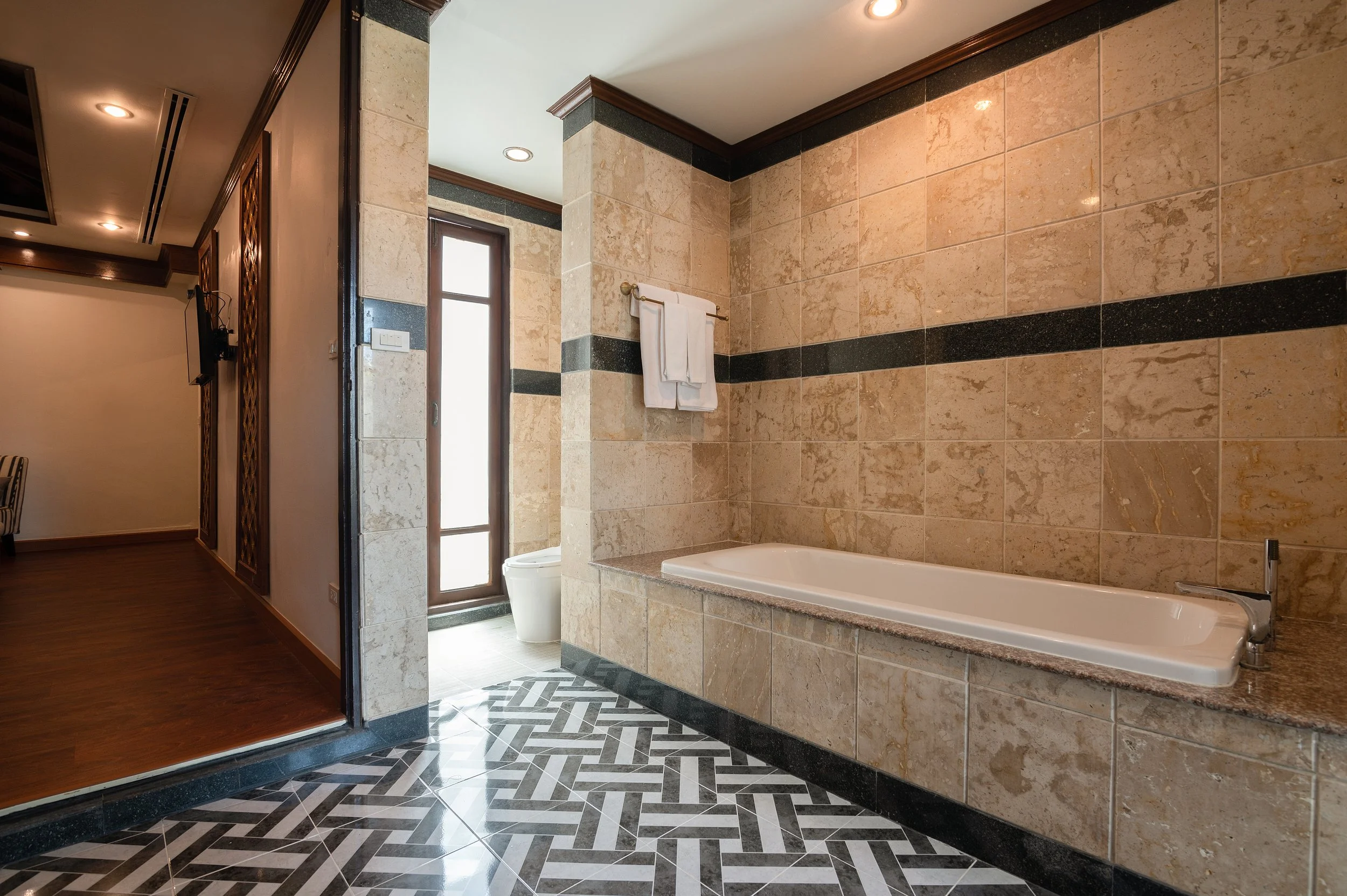 A bathroom with a bathtub on a tiled platform, beige and black wall tiles, a towel rack with folded white towels, a door with frosted glass leading outside, and a patterned black and white floor.