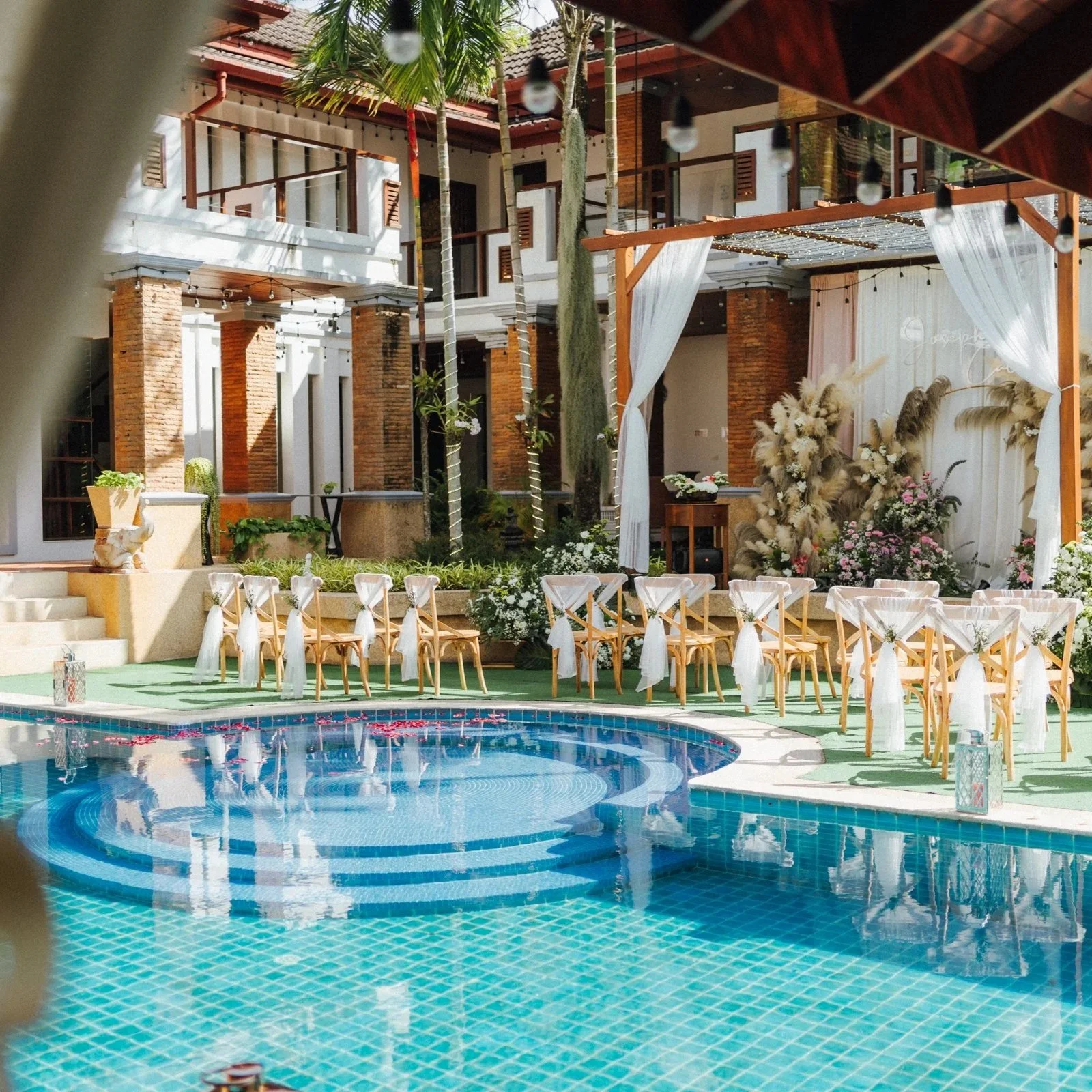 Decorated outdoor poolside area with tables and chairs, set for an event, featuring floral arrangements, string lights, and a backdrop with white curtains.  Pool side party best villa phuket
