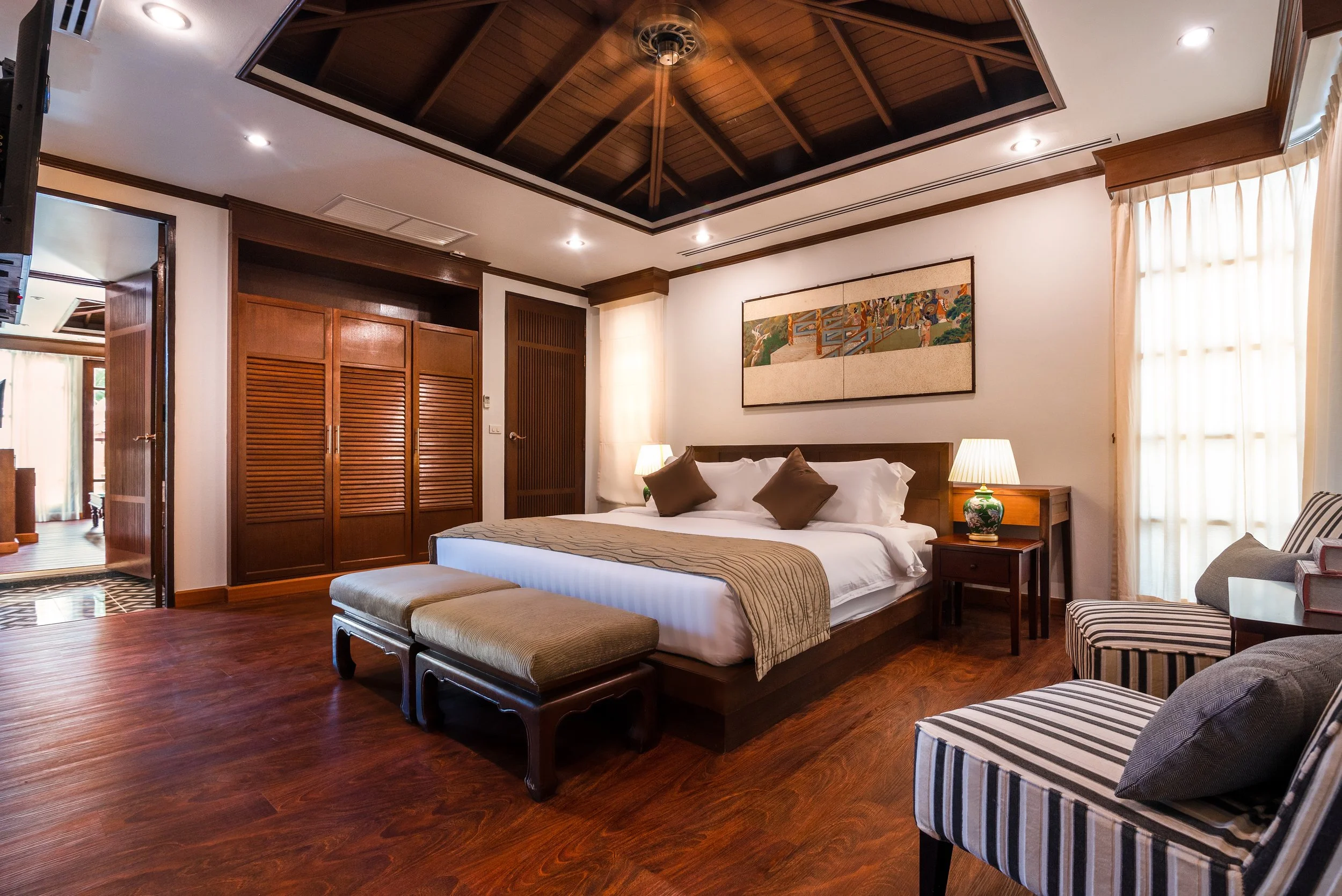 A luxurious room featuring a large bed with white linens pillows, flanked by two wooden bedside tables with lamps. The room has dark wood flooring, a vaulted wood ceiling, and a large window with sheer curtains. There are seating options including st