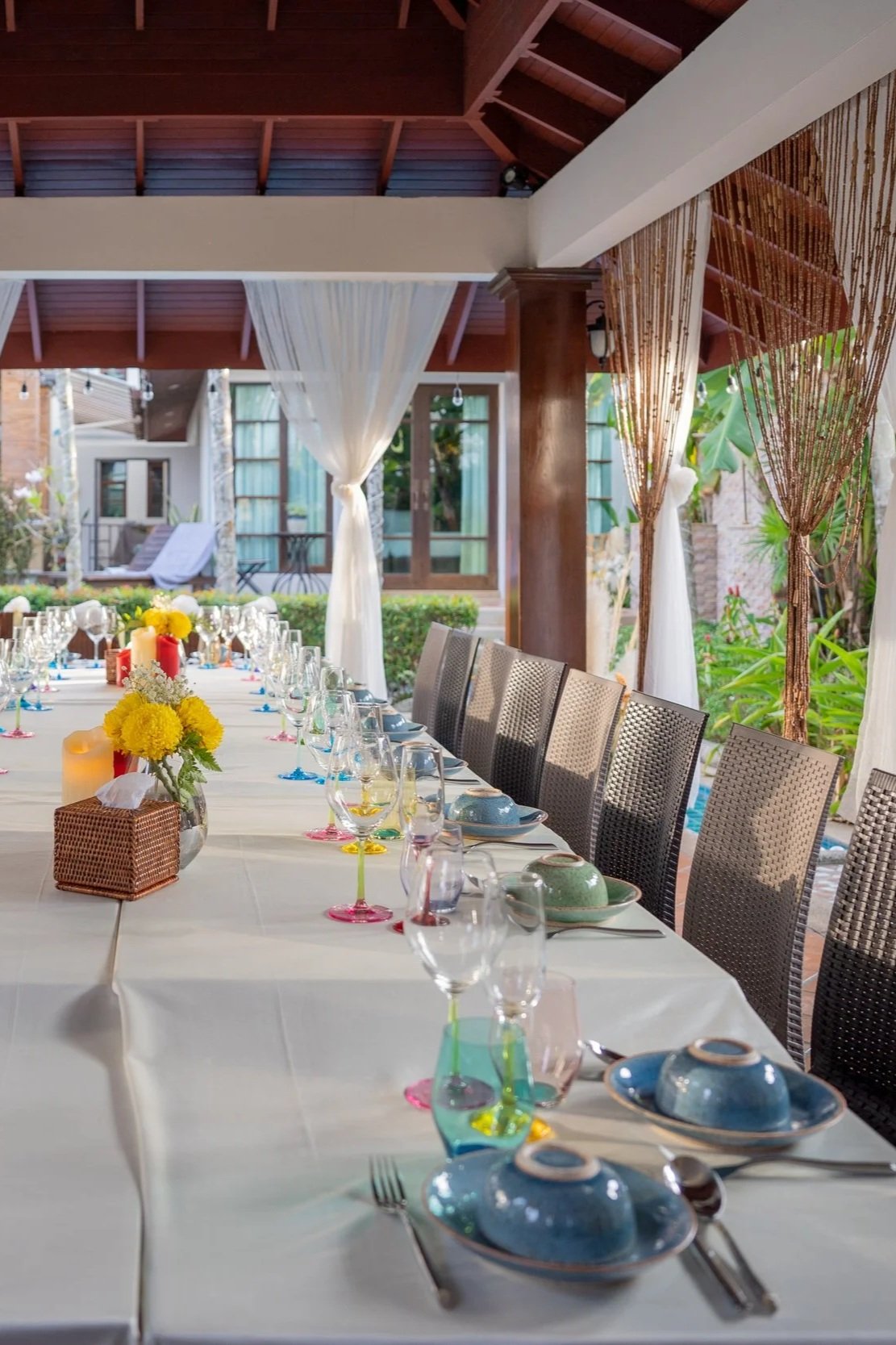 A long dining table set for a meal outdoors, with colorful glassware, plates, and floral centerpieces, under a covered patio with white and brown curtains and surrounding greenery.  Villa private dining Phuket