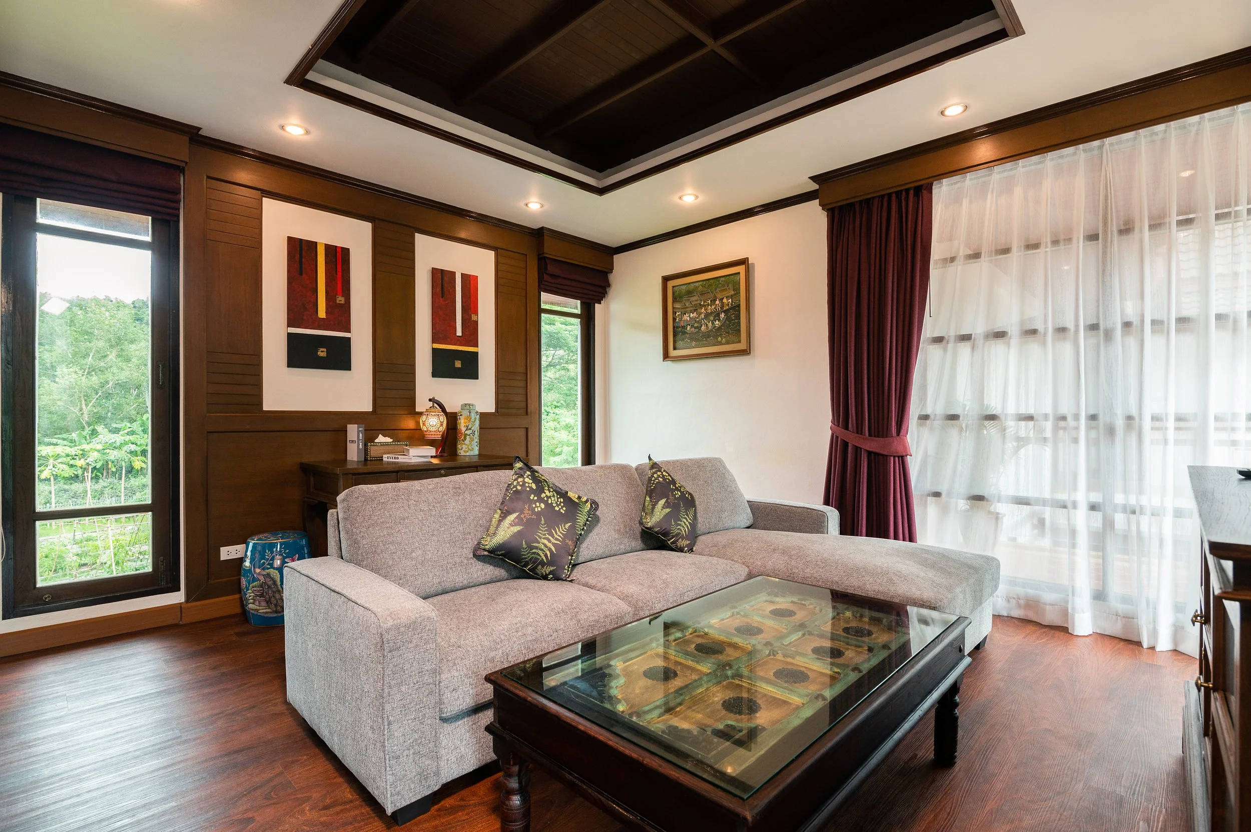 Living room with a gray sectional sofa, decorative pillows, a wooden coffee table with a glass top, dark wood paneling on the walls, colorful artwork, large windows with sheer white curtains and red drapes, hardwood floors, and ceiling recessed light