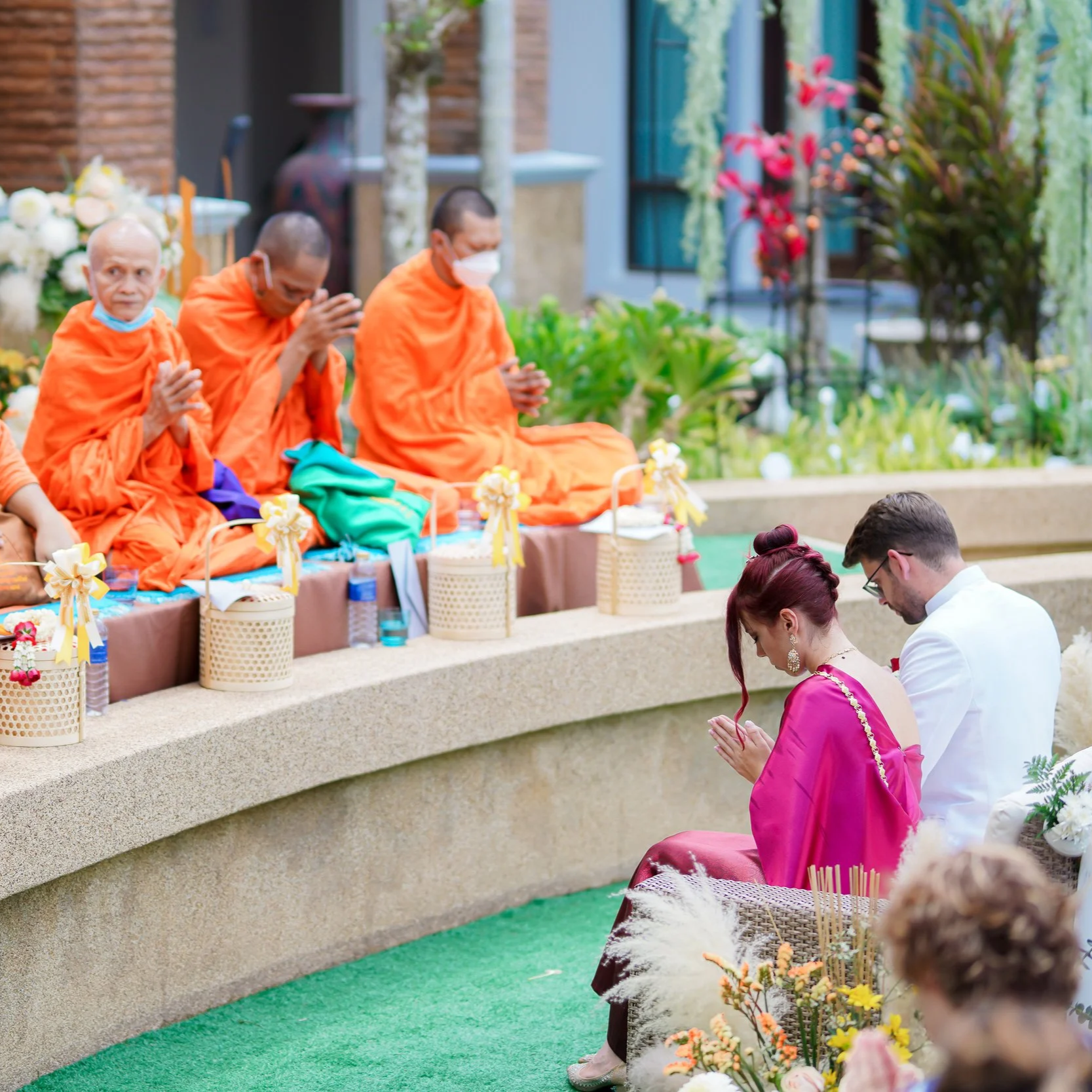People praying at a Buddhist ceremony outdoors with monks in orange robes and laypeople, including a woman in a pink dress and a man in white, kneeling and praying.