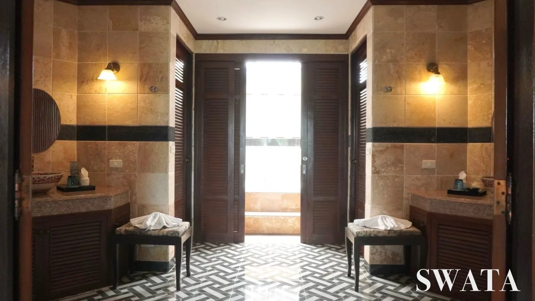 View of an elegant lobby or corridor with tiled flooring, wooden shutters, and warm lighting on the walls. Two chairs with white cloths and side tables are on either side of the open doors, leading to a bright outdoor area.