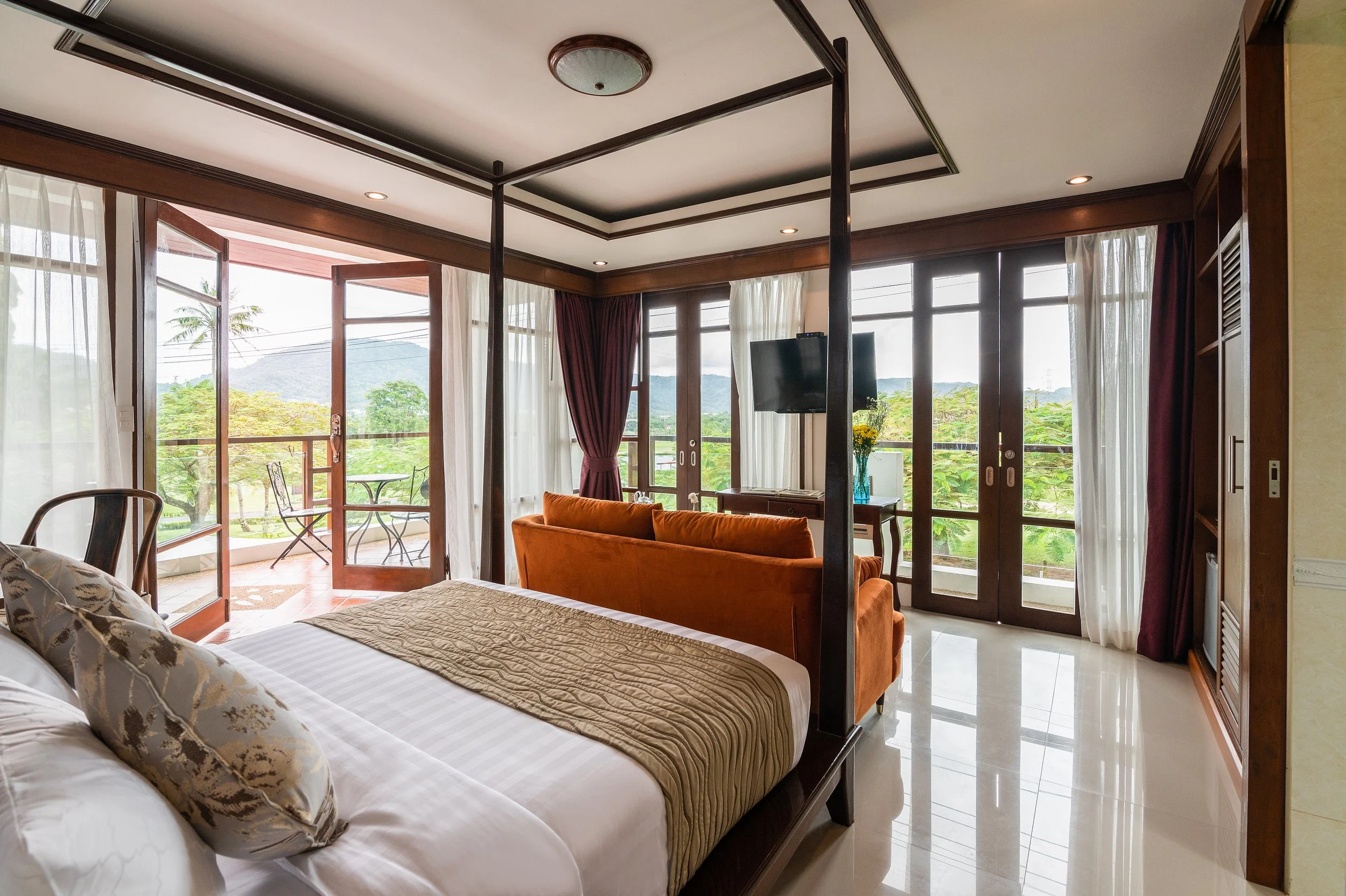 Luxurious hotel room with king-sized bed, orange sofa, flat-screen TV, glass doors leading to balcony with outdoor seating, overlooking green scenery and mountains.