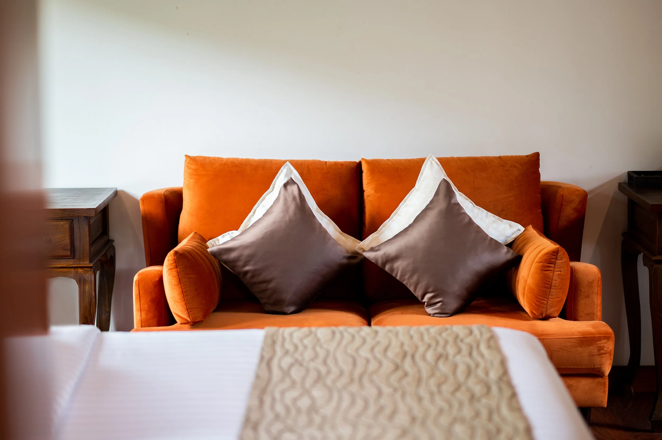 An orange sofa with three pillows, two brown and one cream, in a bedroom with a bed in front and wooden side tables.