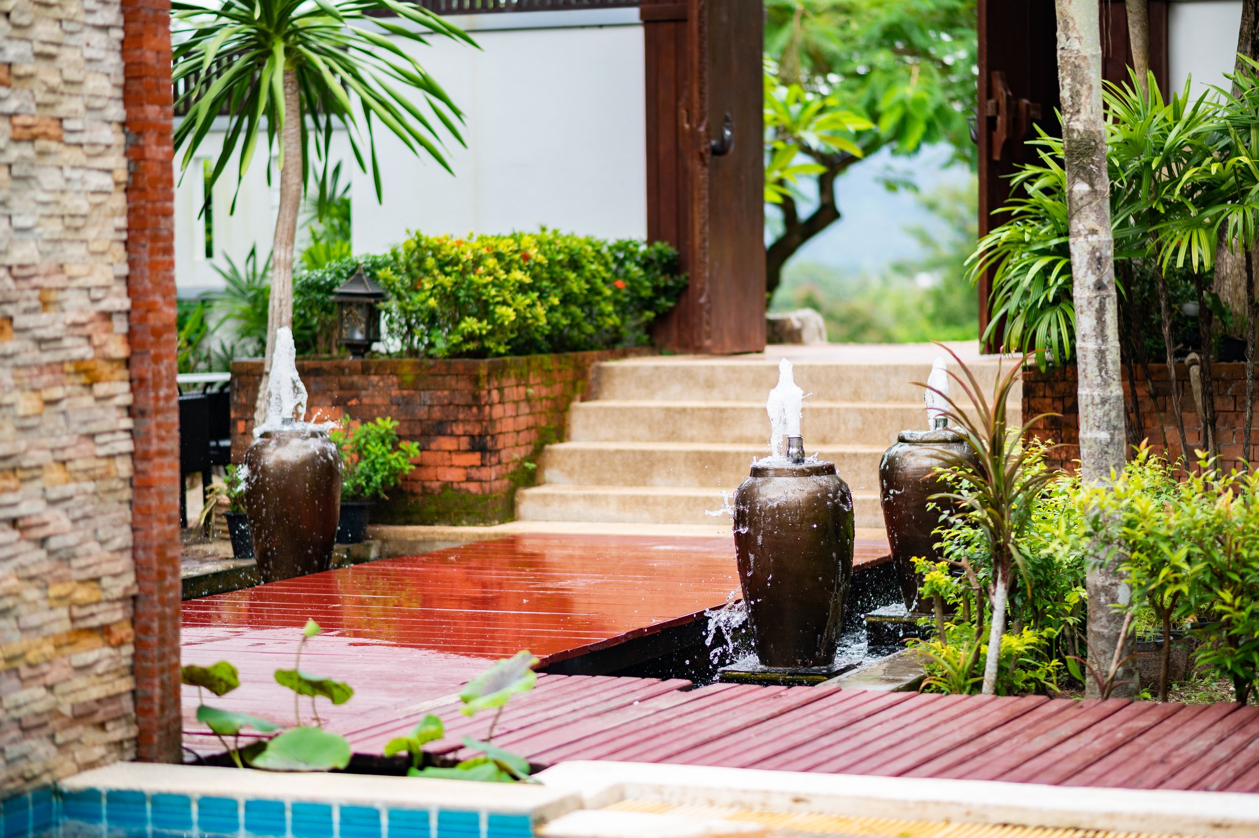 Outdoor garden with water feature, three large brown pots with water fountains, surrounded by lush green plants, trees, and a stone pathway leading to stairs.