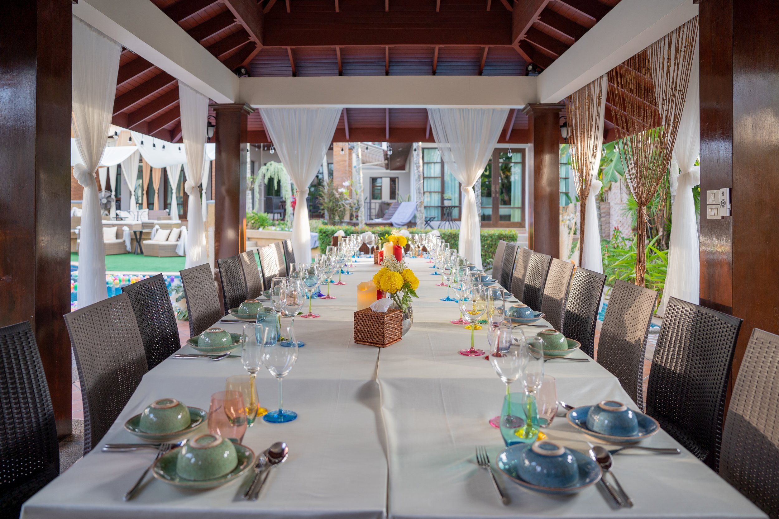 A long dining table set with plates, glasses, cutlery, and yellow flowers in the center, under a wooden pavilion with white curtains, overlooking a garden and pool area.