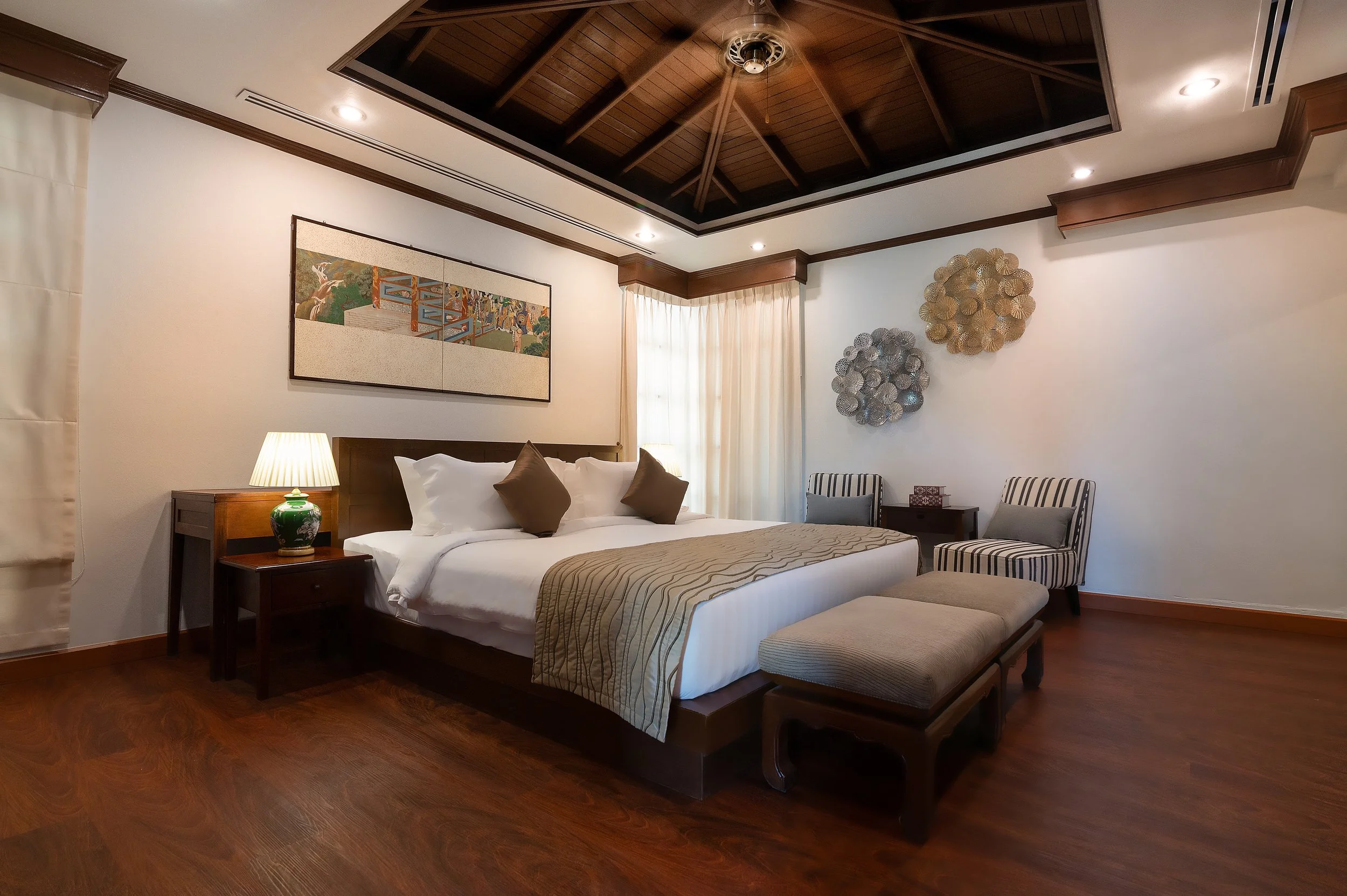 A bedroom with a large bed covered in white linens and brown pillows, a wooden bench at the foot of the bed, two striped armchairs, a wooden side table, and a bedside lamp. The room has wooden floors, a framed artwork above the bed, decorative wall h