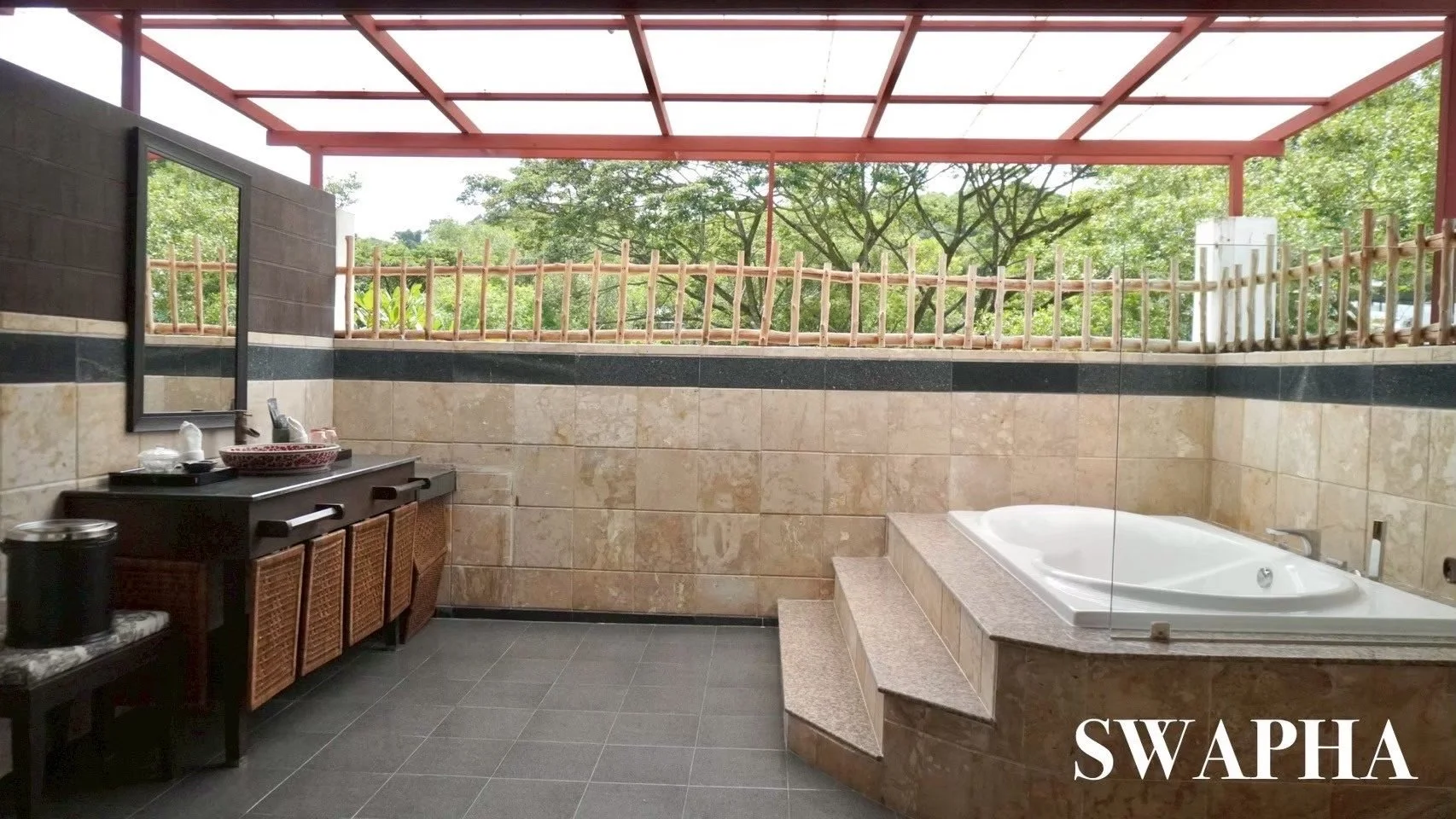 Outdoor bathroom with a jacuzzi tub, a black vanity with a red sink, a mirror, and a bamboo fence, overlooking green trees.