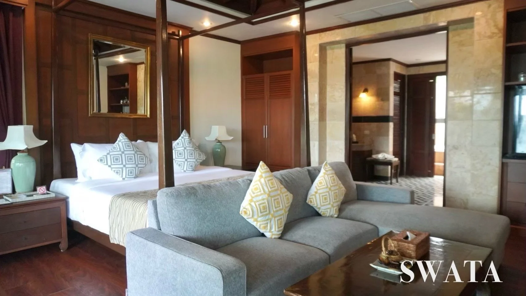 A hotel room with a large bed, decorative pillows, and a wooden headboard. There are two bedside tables with lamps, a gray sofa with yellow-patterned pillows, and a wooden coffee table with a tray and decorative items. The room has wooden wall paneli