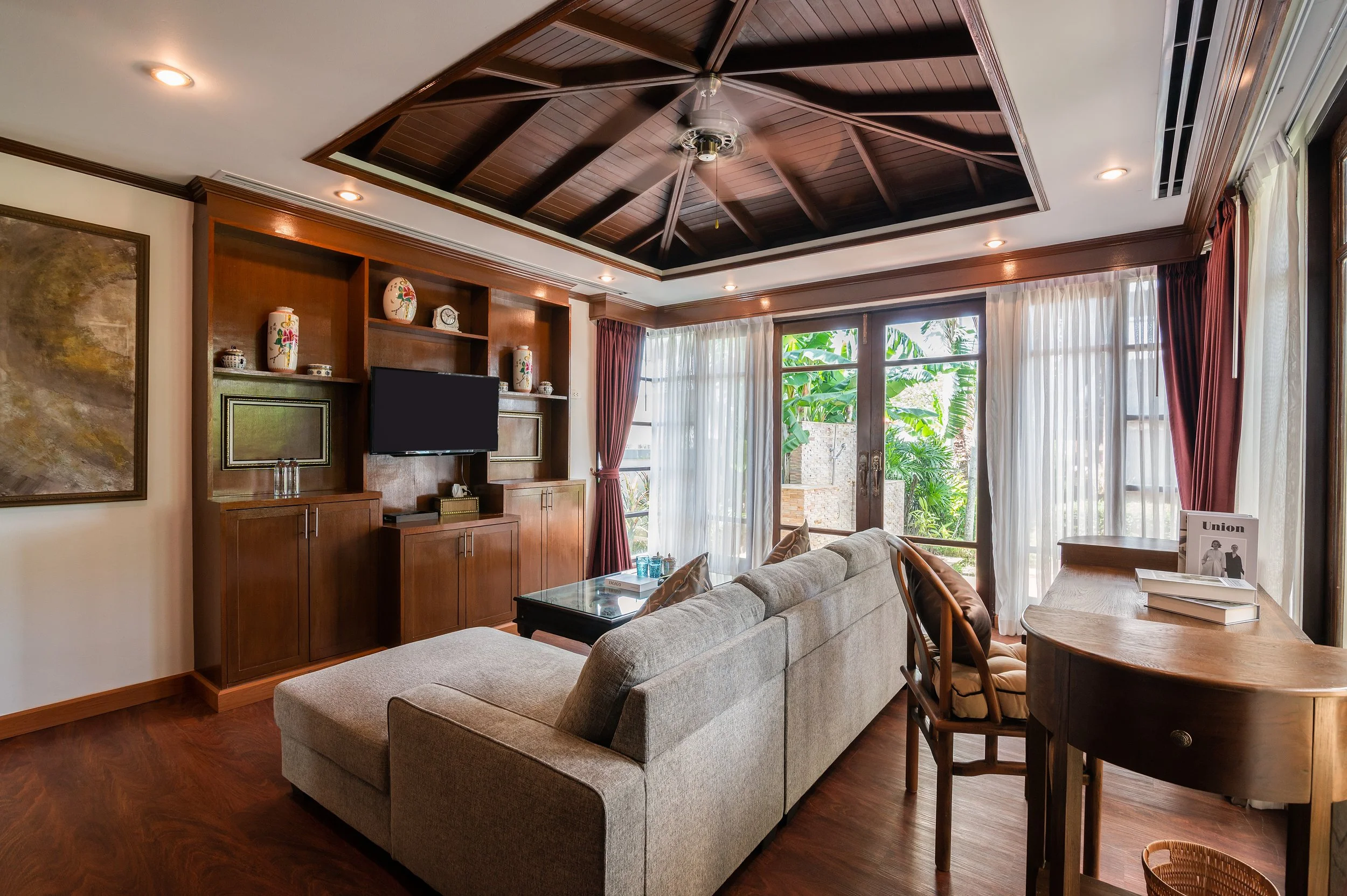 Living room with a beige sectional sofa, a wooden TV cabinet with a mounted flat-screen TV, decorative vases, and a granite coffee table. Large windows with white sheer curtains and red drapes overlook greenery outside. The ceiling has wooden panelin