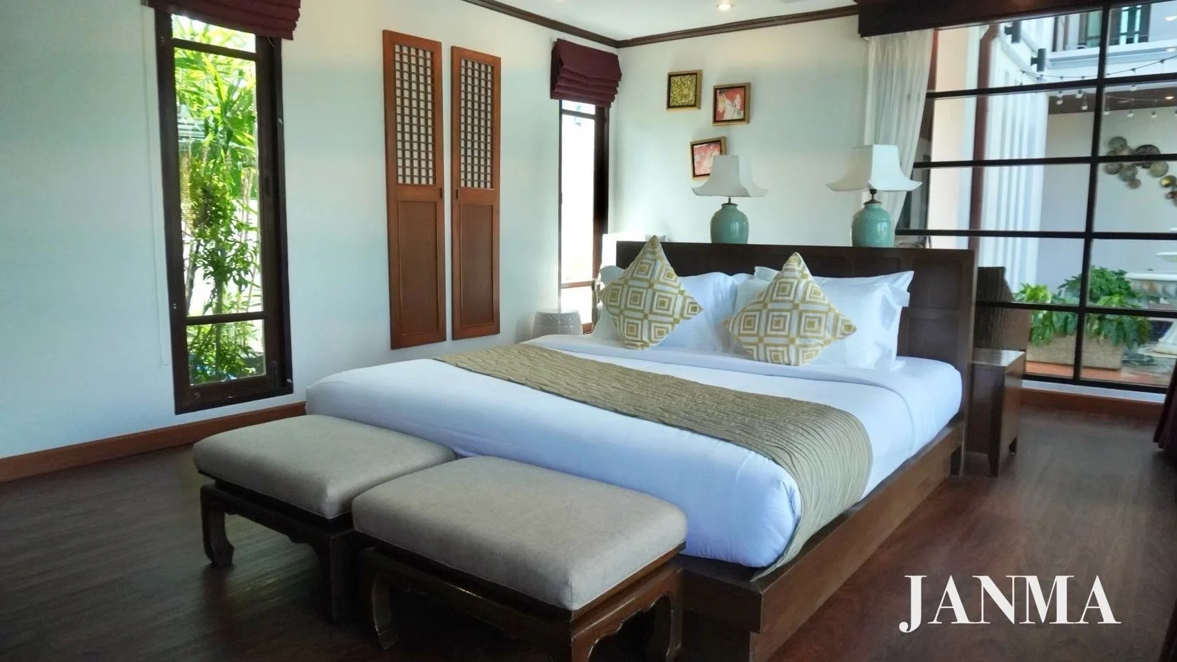 A cozy bedroom with a large bed featuring white bedding, decorative yellow and white pillows, and a gold bed runner. There are two matching bedside lamps on teal vases behind the bed, wooden framed windows with curtains, wall art, and a bench at the 