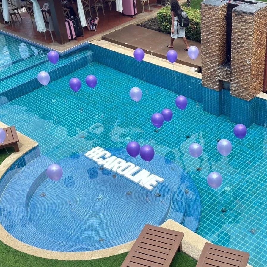 View of a swimming pool with purple and lavender balloons floating on the water, a large white hashtag sign reading '#CAROLINE' at the bottom, and a person walking nearby with a backpack.  Birthday party celebration  best villa phuket