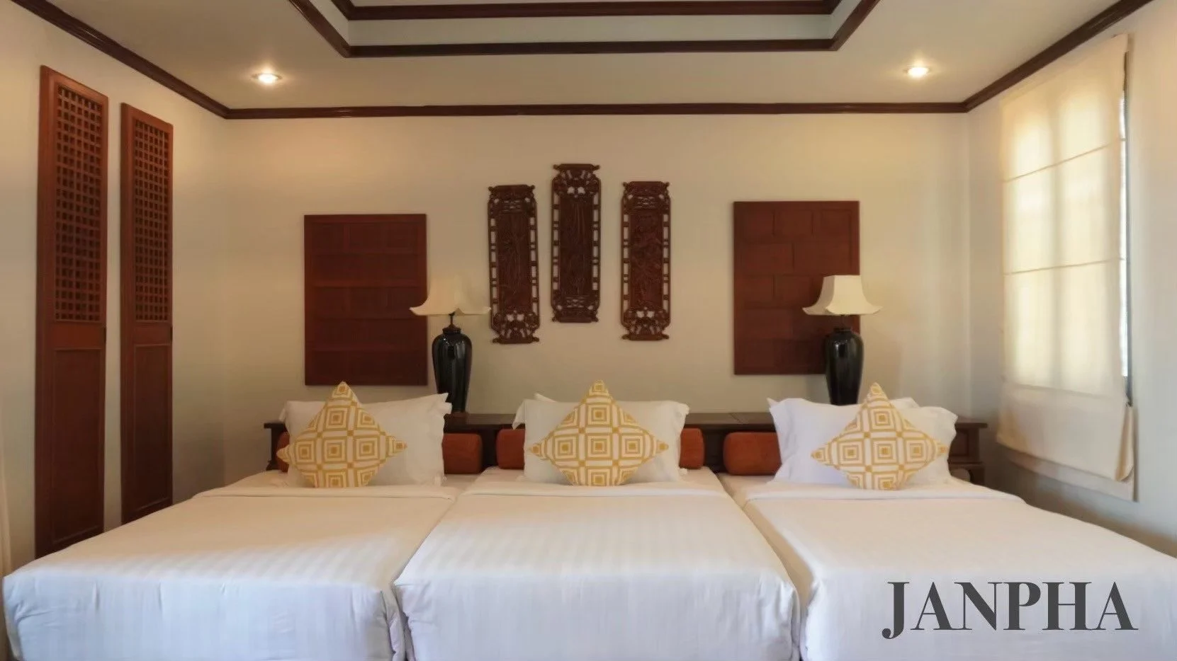 A bedroom with three beds, each with white linens and yellow patterned throw pillows, a dark wooden headboard, two bedside lamps with black bases and white shades, decorative wooden wall art above the beds, wooden window coverings, and the word 'JANP