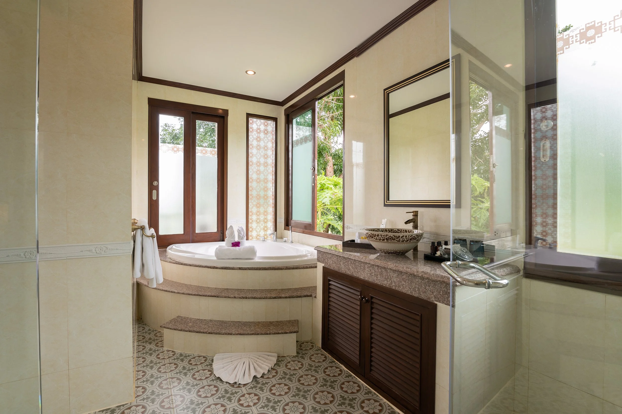 A luxurious bathroom with a large bathtub near glass doors and windows, a granite countertop with a stylish basin sink, and decorative tile flooring.
