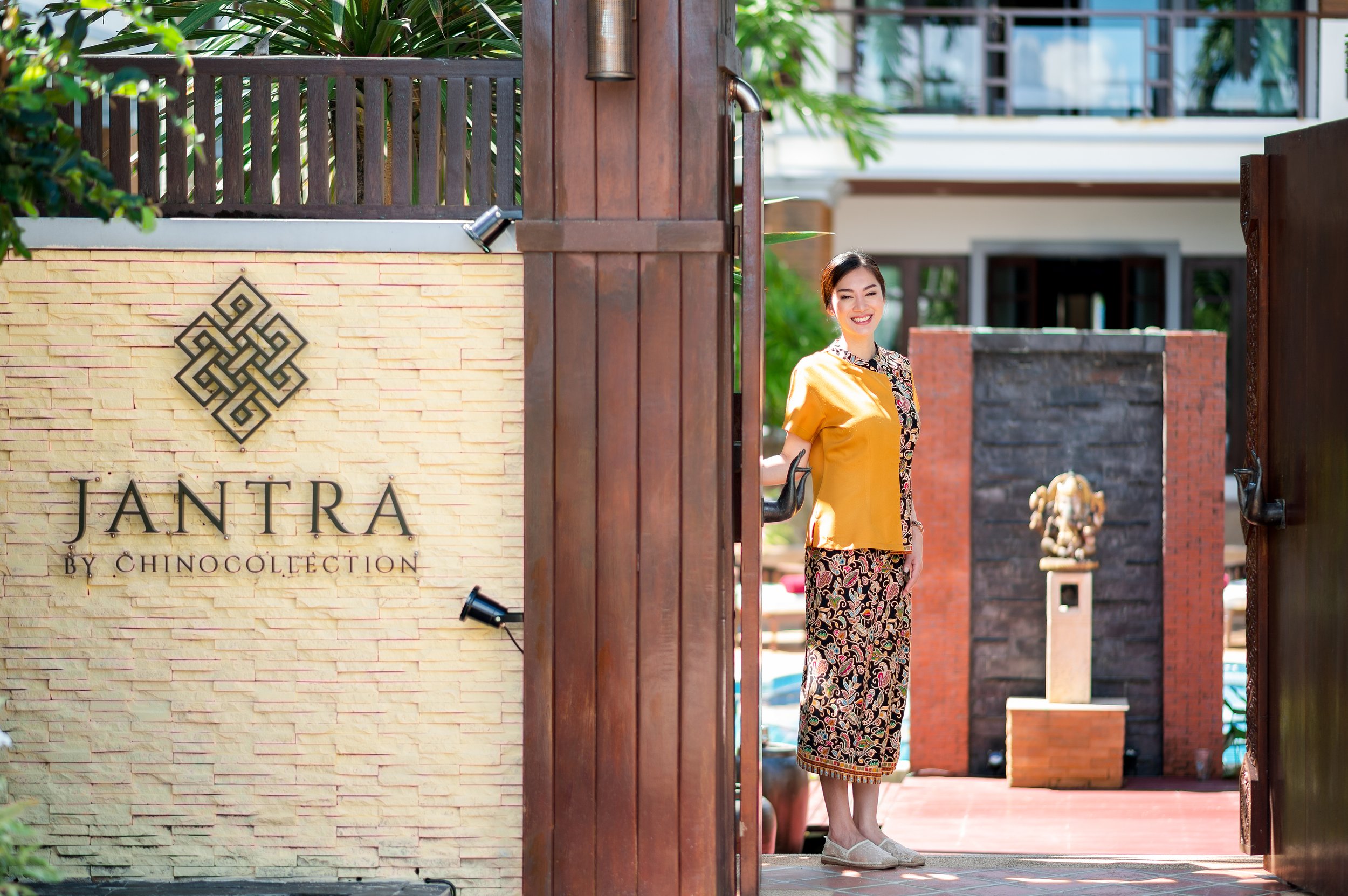 A woman dressed in traditional clothing stands at an open gate, smiling. The gate has a sign that reads 'JANTRA BY CHINOCOLLECTION' and is surrounded by lush greenery and a modern building in the background. Best large group villa