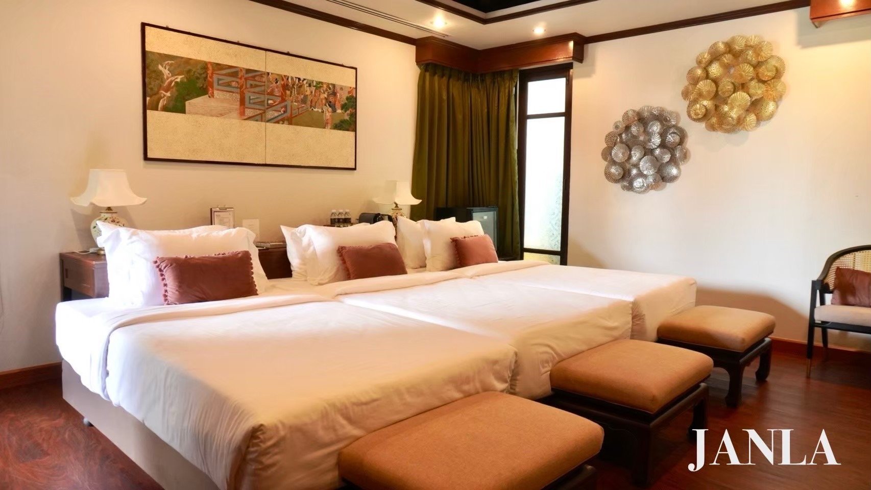 A hotel room with a large bed, multiple pillows, bedside lamps, a framed artwork above, and decorative wall hangings, with a small window and green curtains.