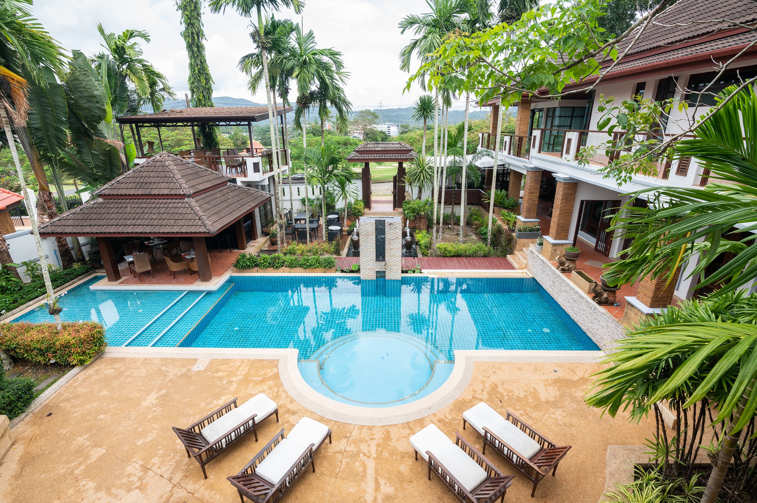A luxurious outdoor swimming pool area with lounge chairs, surrounded by lush green tropical trees and plants. There are multiple buildings with balconies and a covered pavilion with seating. The scene is serene with a backdrop of hills and cloudy sk
