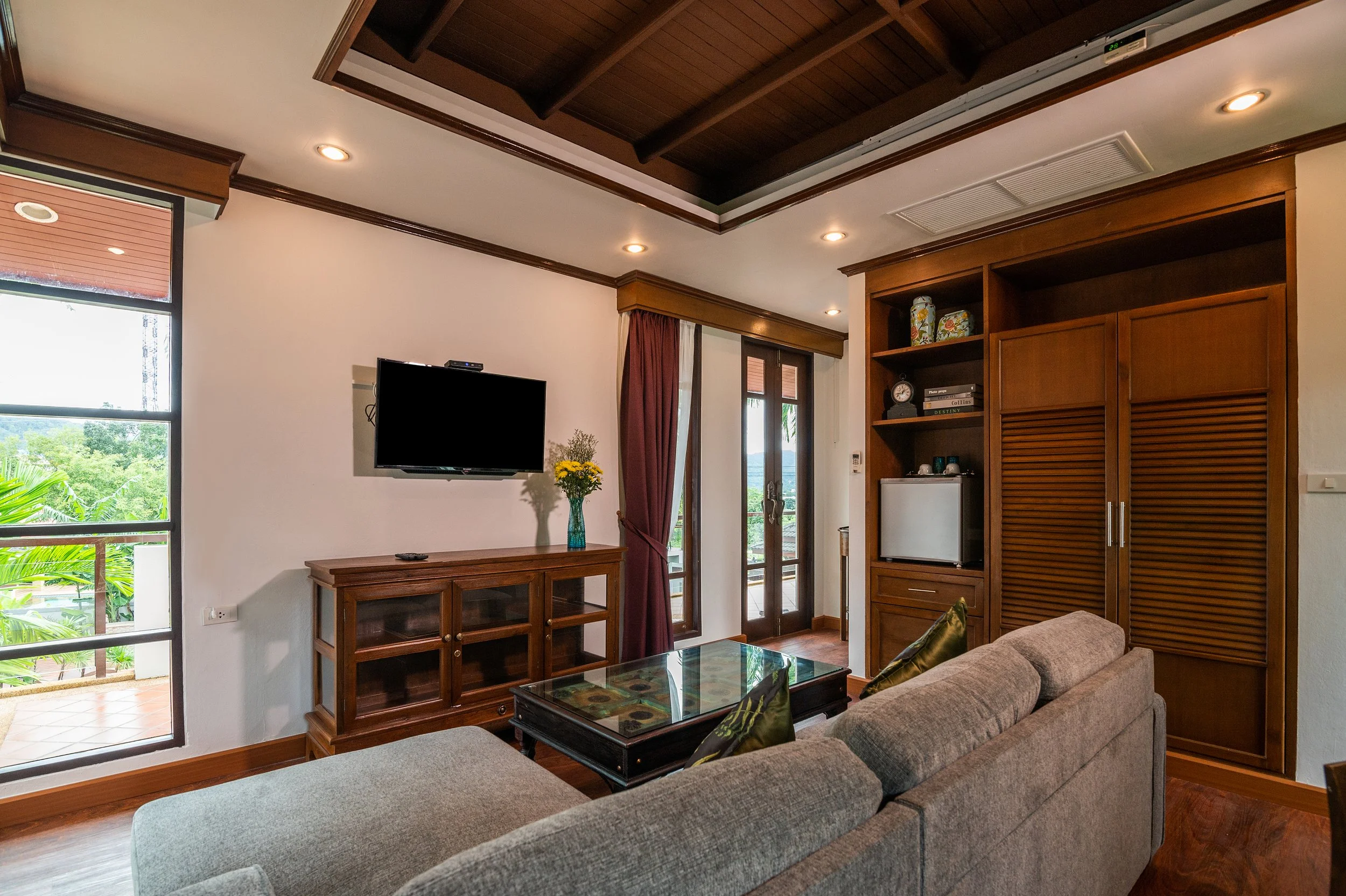 Living room with a wall-mounted TV, wooden entertainment center, glass coffee table, L-shaped sofa, shelves with decorative items, and a balcony with glass doors showing greenery outside.