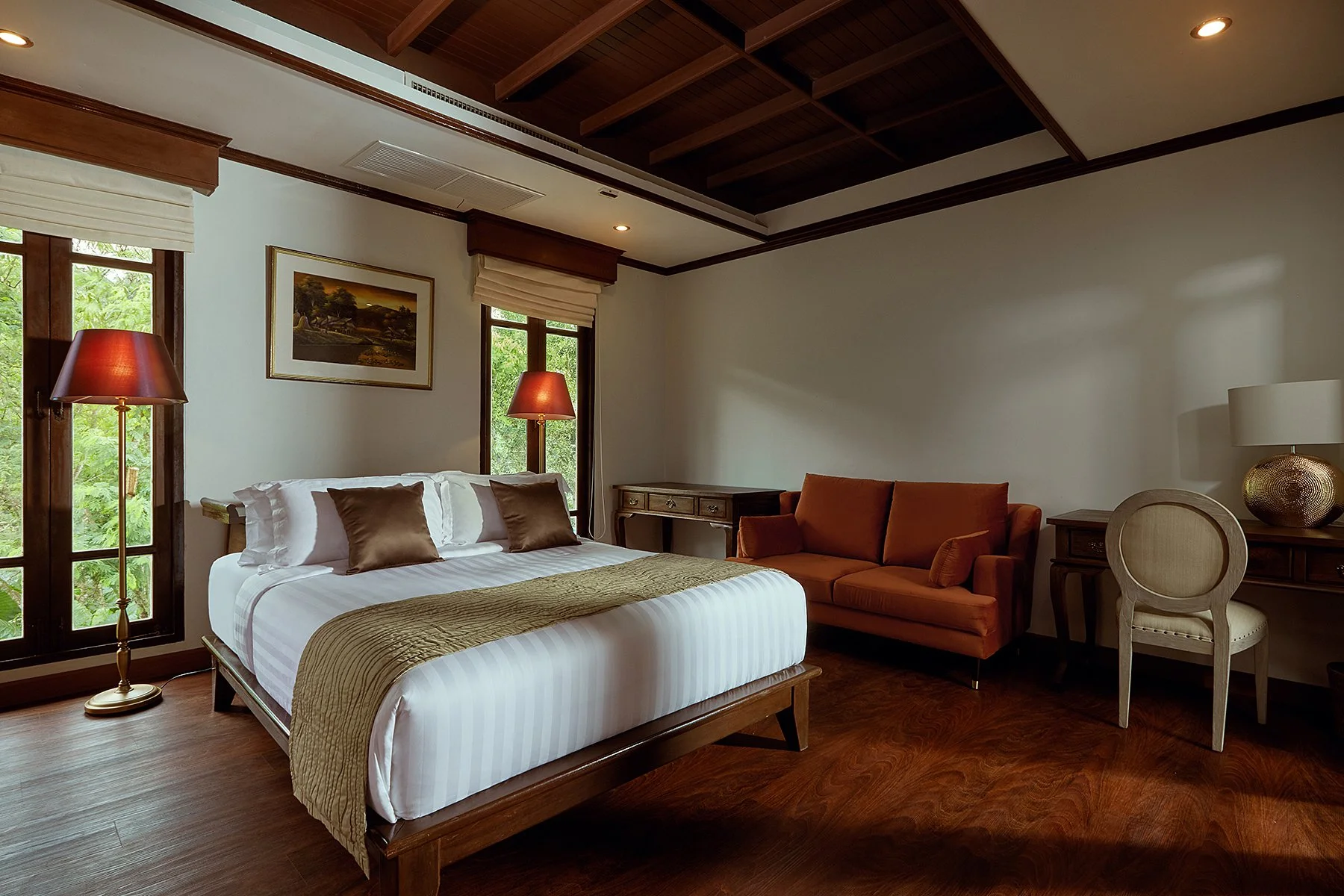 A cozy bedroom featuring a large bed with white linens and brown pillows, beside a floor lamp. There is a window with greenery outside, a small desk, and a burnt orange sofa in the corner. A wooden dresser with a lamp is on the right, and the ceiling