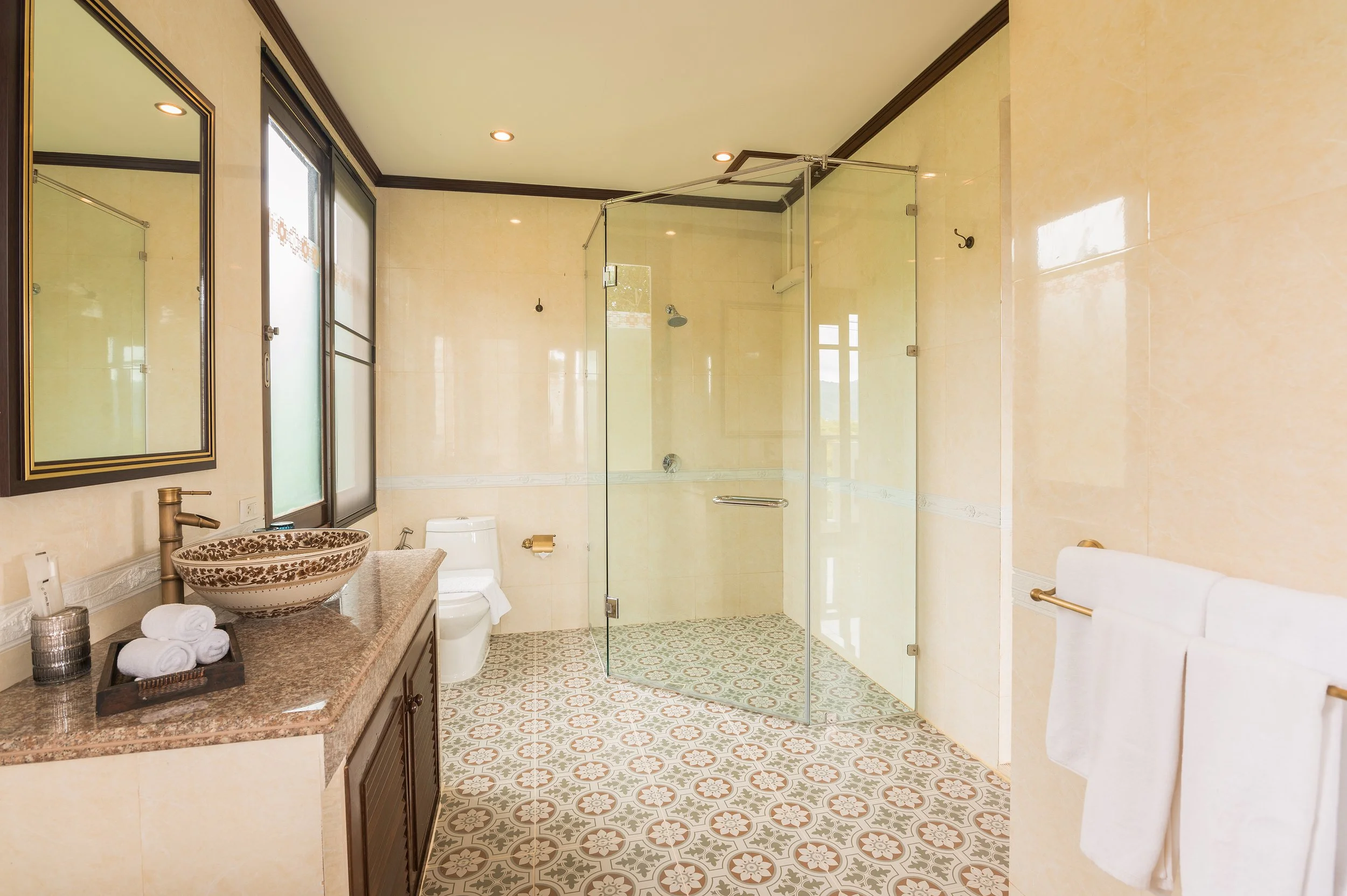 Bathroom with beige tiled walls and patterned floor, glass shower enclosure, toilet, wooden vanity with stone countertop, decorative sink, mirror, towel rack with white towels, window with frosted glass, and ceiling lights.