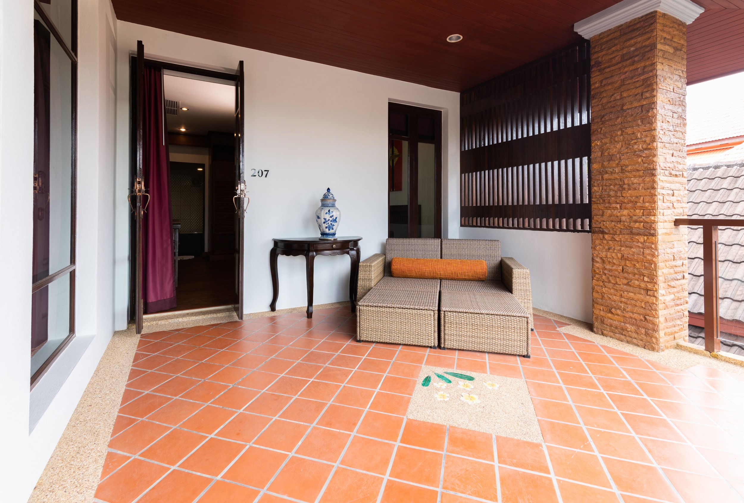 Outdoor balcony with orange tile floor, beige woven sofa with orange cushion, black wooden table with blue and white vase, white walls, brick and wood accents, and glass door with purple curtains.