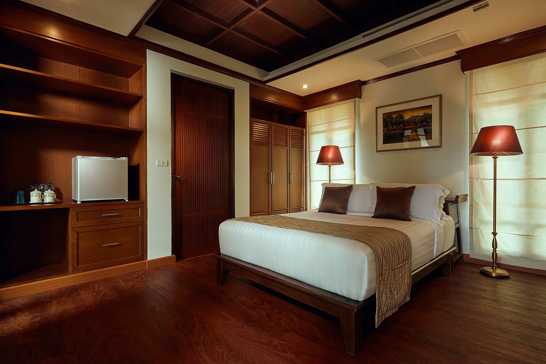 A hotel room with a bed, two lamps, wooden furniture, and a window with beige curtains