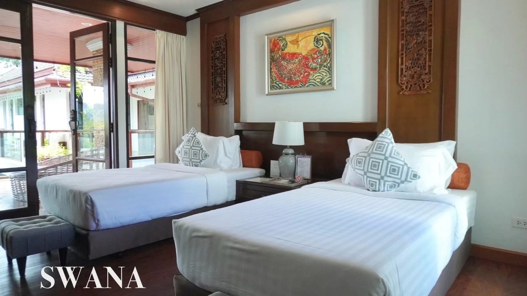 Two single beds in a hotel room with white linens, decorative pillows, a bedside table with a lamp, and a painting above the headboard. There is a sliding glass door leading to a balcony.