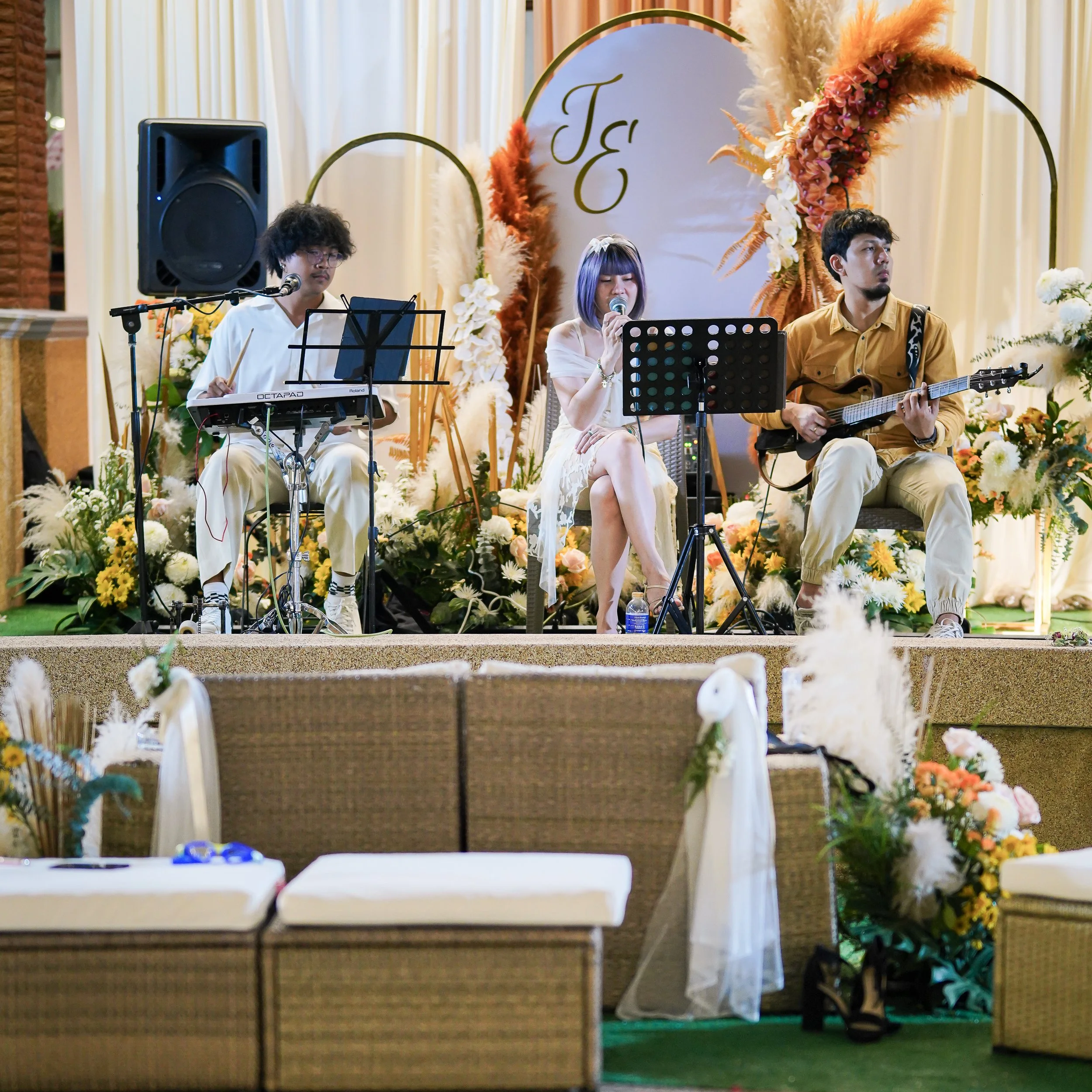 Three musicians perform on a decorated stage surrounded by flower arrangements at an event, with a woman singing into a microphone in the center, flanked by two male guitarists.  Party celebration best villa phuket. Birthday, anniversary