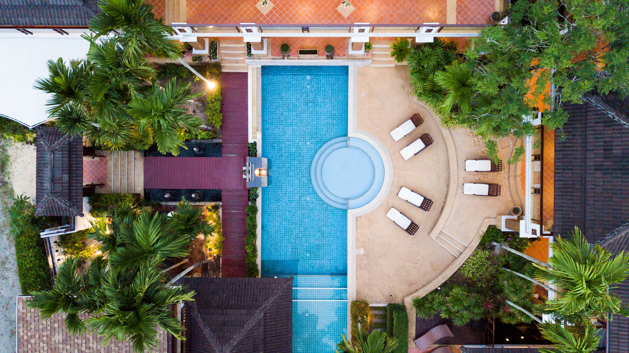 Top-down view of a swimming pool area with lounge chairs, surrounded by palm trees and landscaped greenery at a resort or hotel.