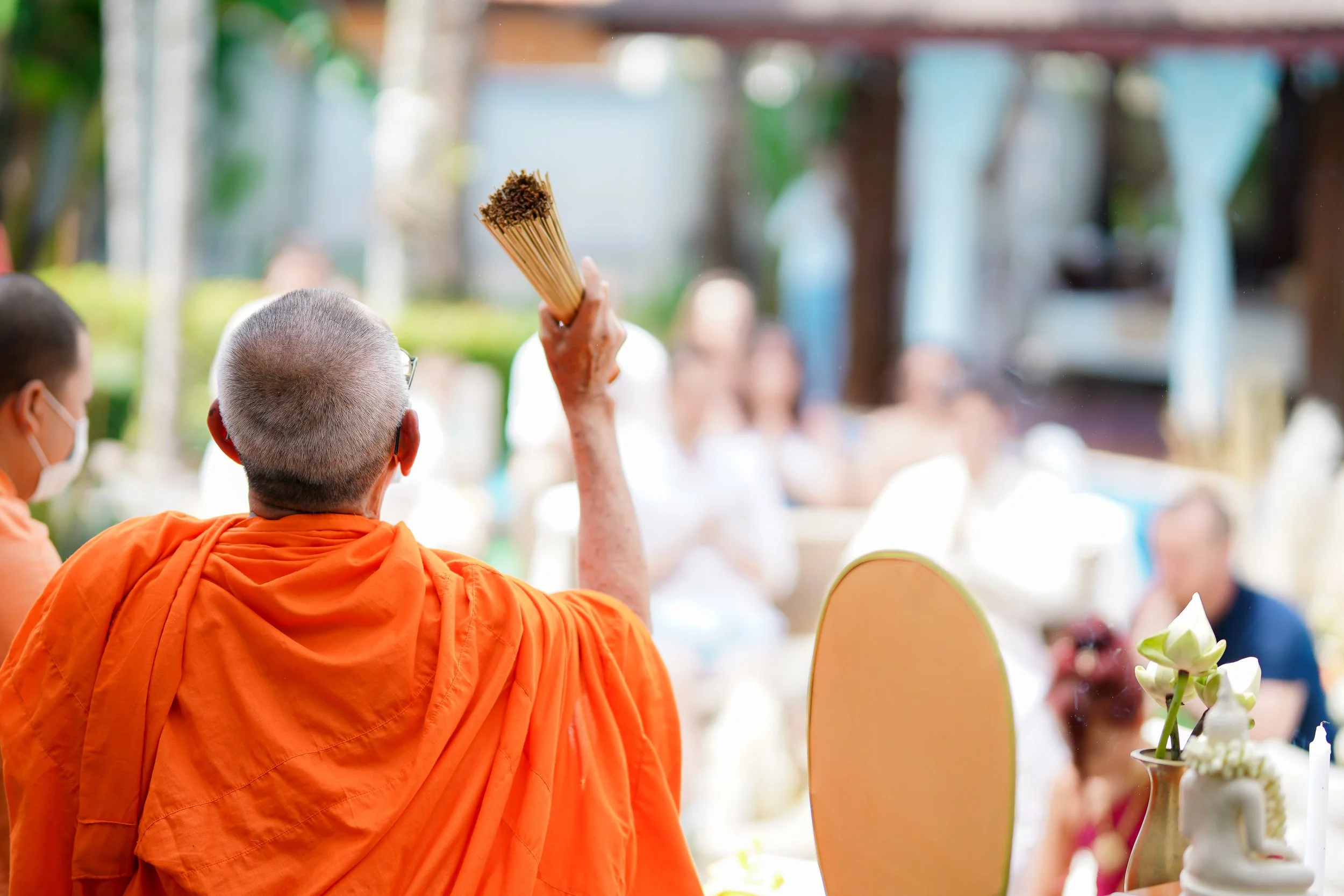 A Buddhist monk in orange robes raising a stick in a ceremony, with an outdoor gathering of people in the background, some wearing masks.