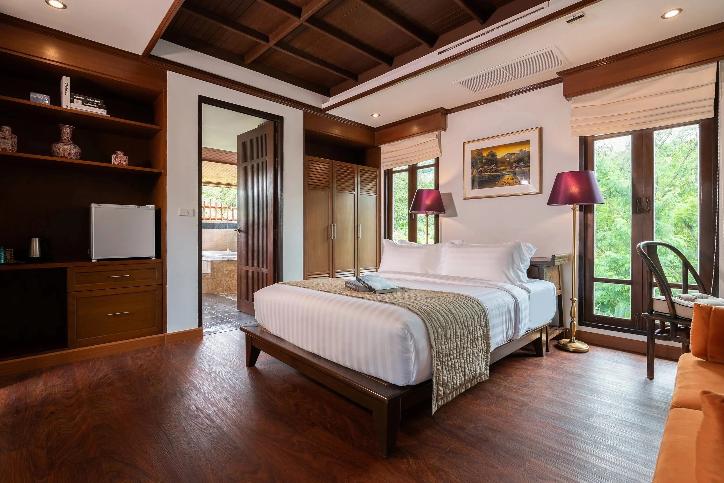 A cozy hotel room with a large bed, wooden furniture, two purple lamps, and a view of green trees through the windows.  Thai style best villa Phuket