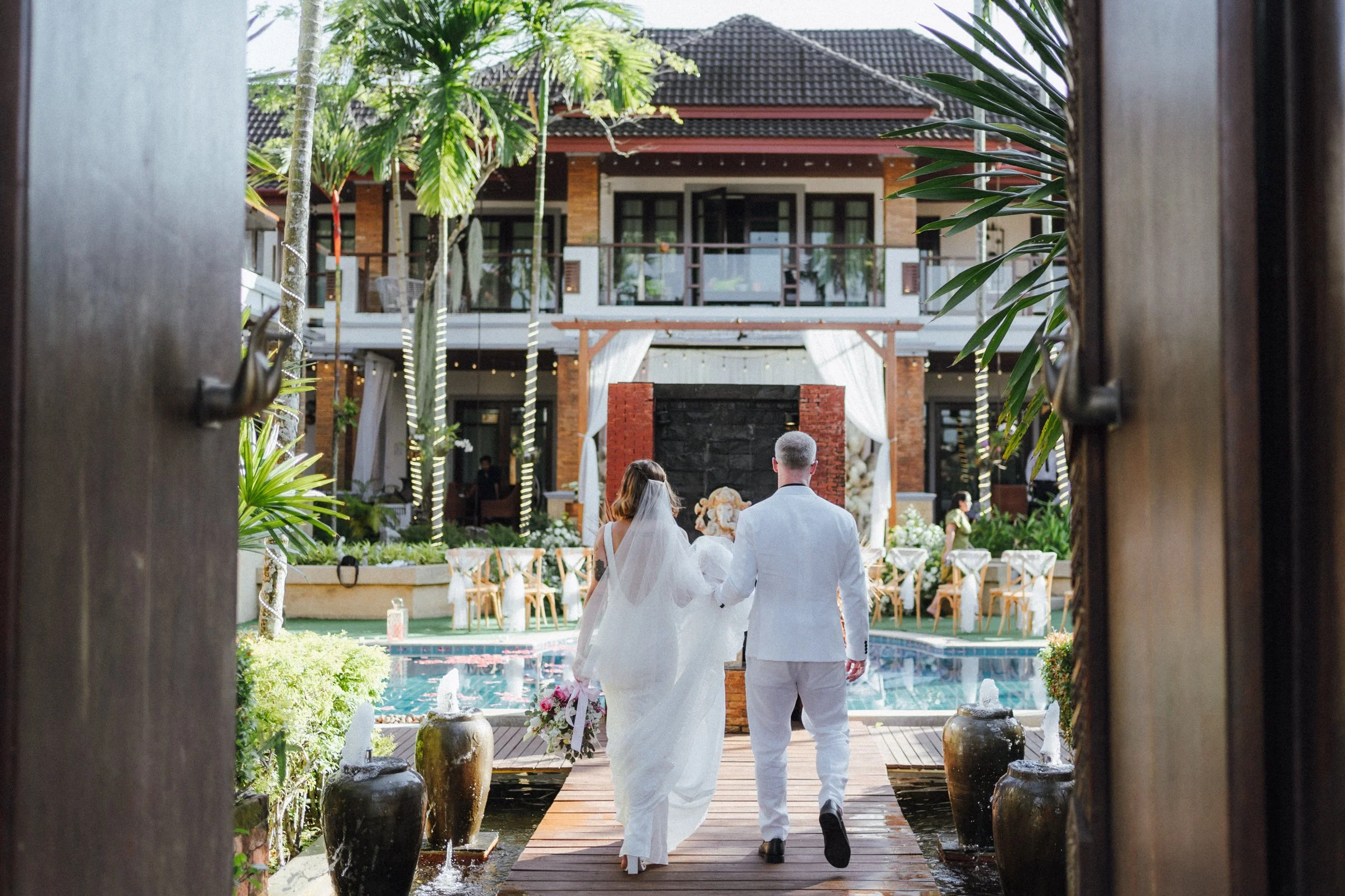 A bride and groom walking hand in hand at an outdoor wedding venue, with a pool, chairs, and a large house in the background. Luxury phuket wedding in large group best villa