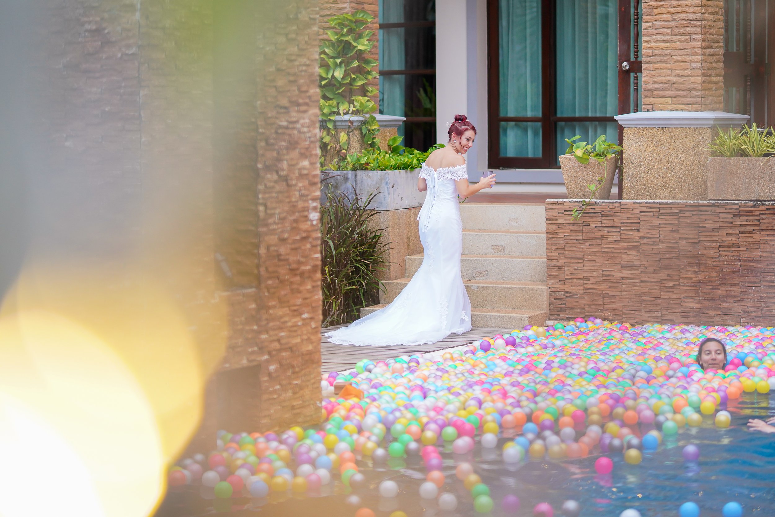 A woman in a white wedding dress standing on steps near a pool filled with colorful plastic balls, smiling and holding a glass, while another woman swims in the pool with only her head visible.
