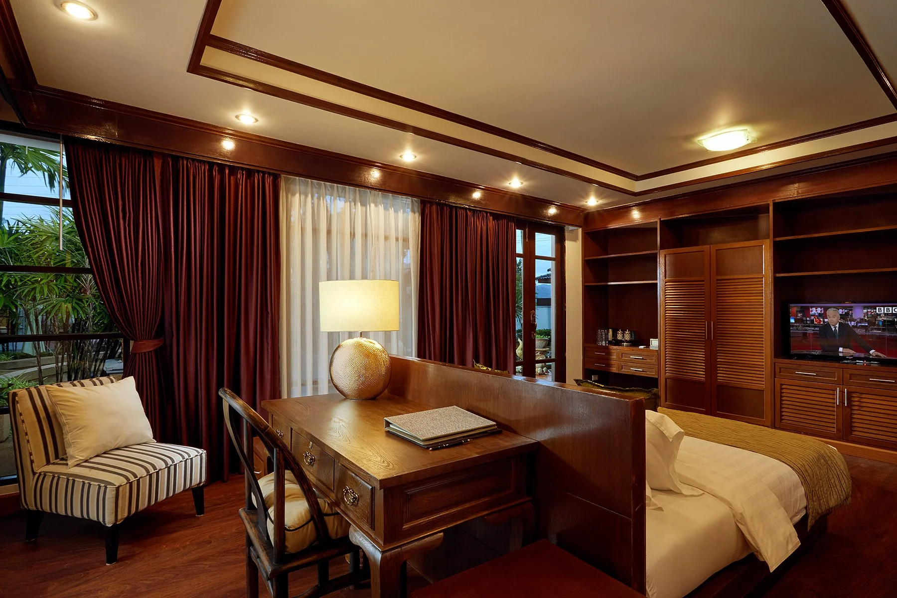 A cozy hotel room with a bed, wooden furniture, a lamp, a striped armchair, and a large window with maroon curtains, showcasing greenery outside.