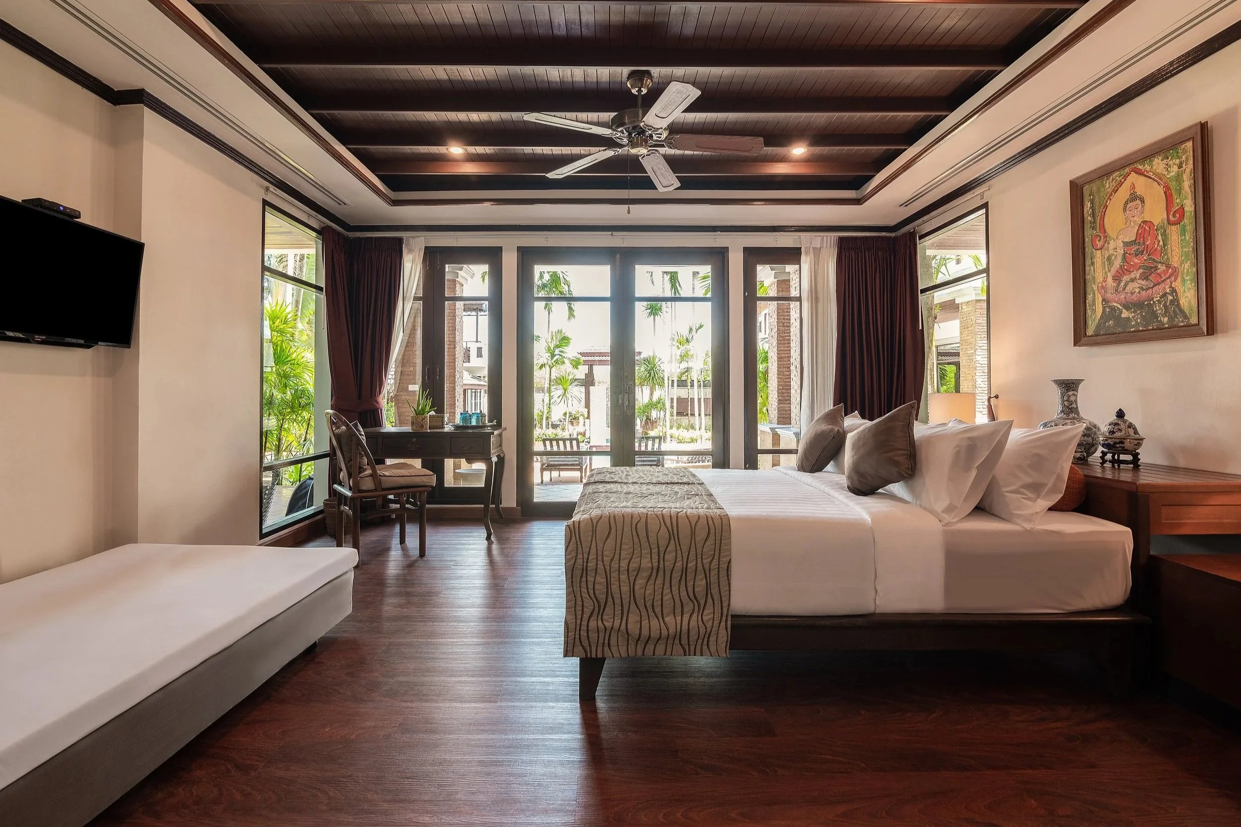 Interior of a villa room featuring a large bed with pillows, a small sitting area with a desk and chair, a TV mounted on the wall, and glass sliding doors leading to a balcony with views of palm trees and neighboring buildings.  Best large group