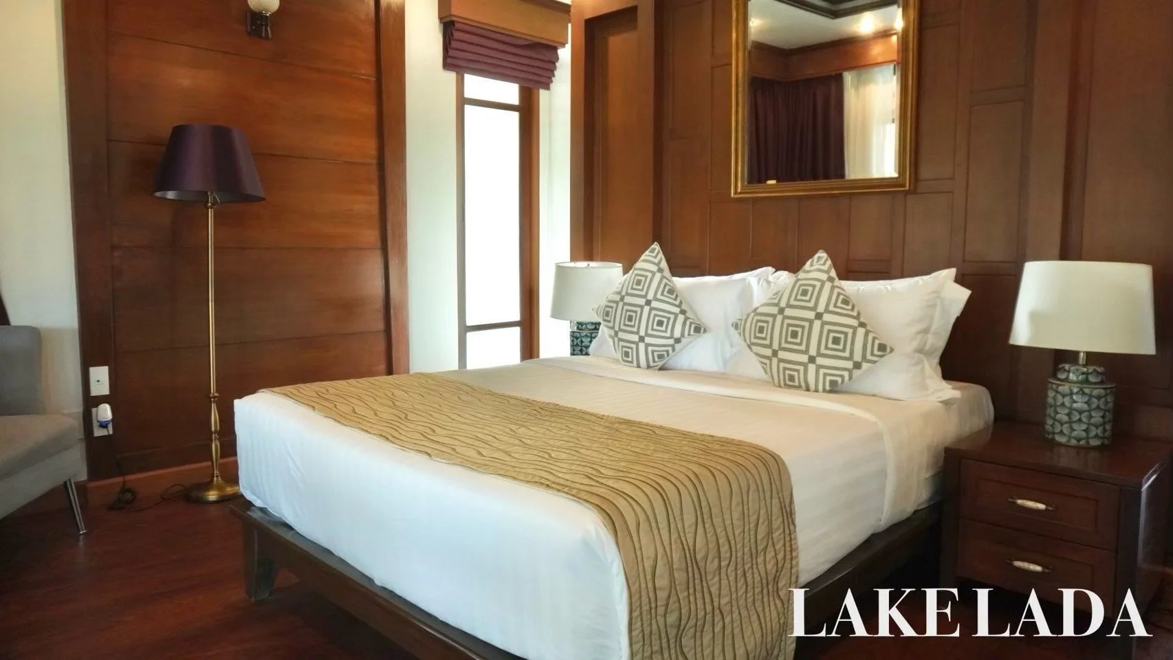 A hotel room with a neatly made bed, wooden walls, and bedside lamps. There are decorative pillows on the bed, a lamp on a nightstand, and a mirror on the wall.