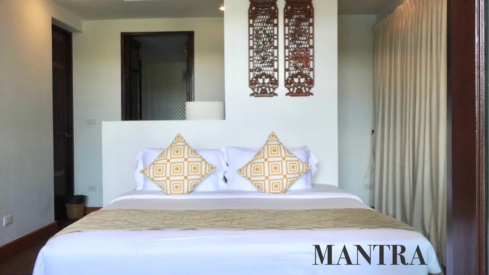 A neatly made bed with white sheets and two yellow patterned pillows in a room with white walls, wooden accents, and decorative wall art.